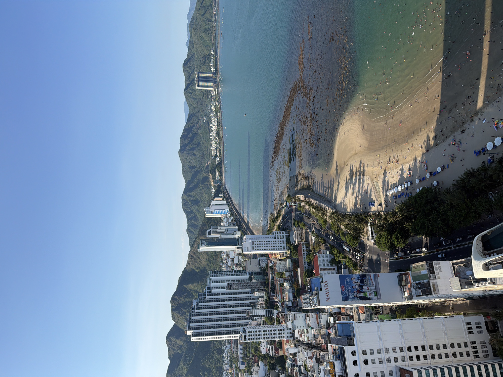 An aerial drone shot captures a bustling beach scene and urban landscape in Bac Nha Trang Ward, Vietnam, under a clear blue sky. A crescent-shaped sandy beach, populated with numerous sunbathers and swimmers, stretches along the turquoise waters of the sea. Umbrellas dot the sand, providing shade to beachgoers. Offshore, rocky formations emerge from the shallow water. The coastline is lined with a wide road bordered by palm trees, with a steady stream of cars and motorcycles. Adjacent to the road, a dense urban environment rises, dominated by a cluster of modern high-rise buildings, primarily white and grey, some featuring intricate architectural designs. One prominent building displays a large vertical billboard advertising "NAVADA" and a beer brand. In the background, lush green mountains form a scenic backdrop to the city and coastline. The image conveys a vibrant atmosphere of a popular tourist destination, blending urban development with natural coastal beauty.