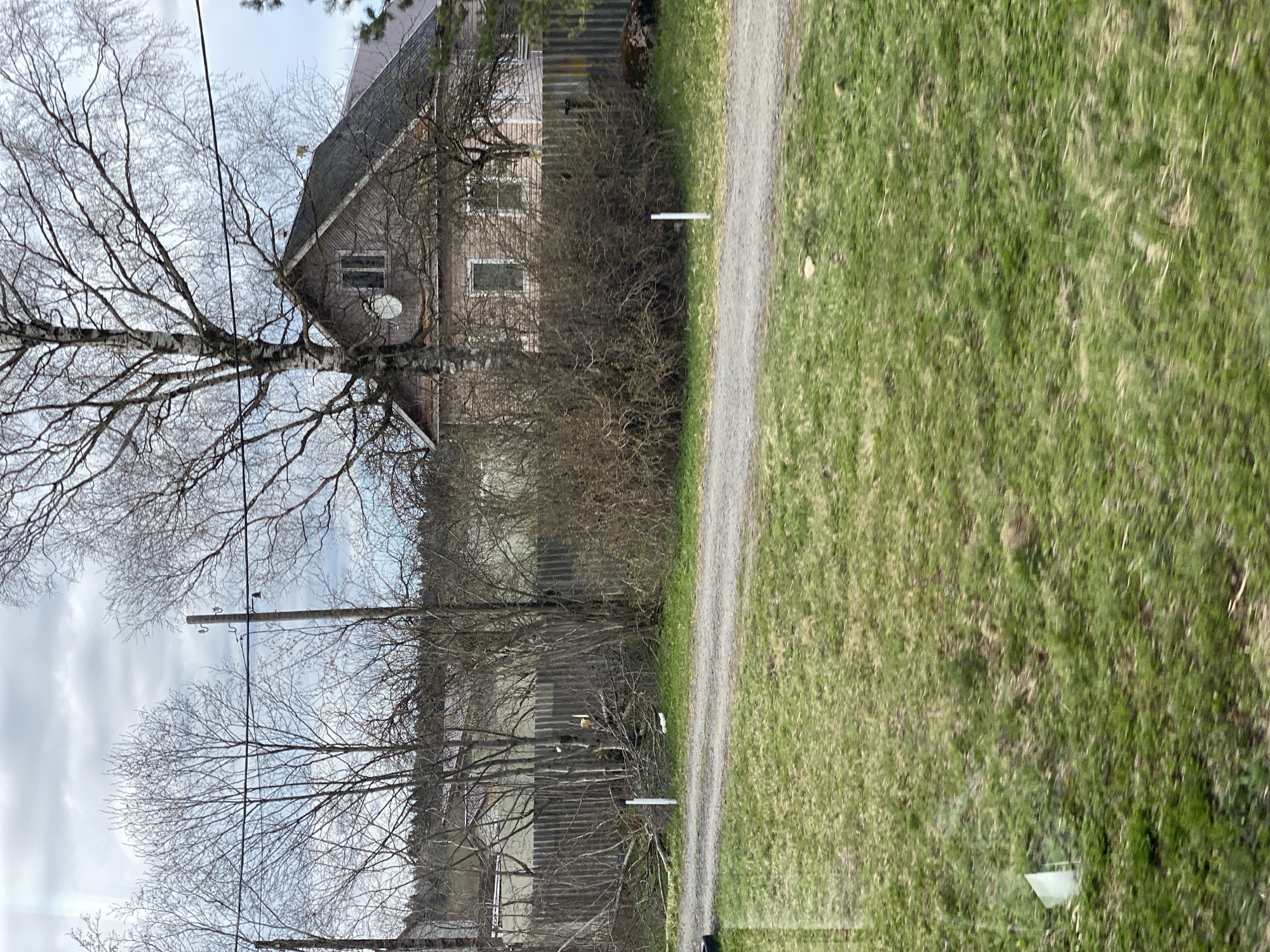 The image shows a rural scene. A dirt road runs through the center of the frame, flanked by a green grassy area on the right. On the left, there is a house with a gray roof and siding, partially obscured by bare trees and bushes. A telephone pole with wires runs vertically through the left side of the image. The sky is overcast. The location is potentially in Kas'kovo, Russia, although this cannot be confirmed from the image alone.