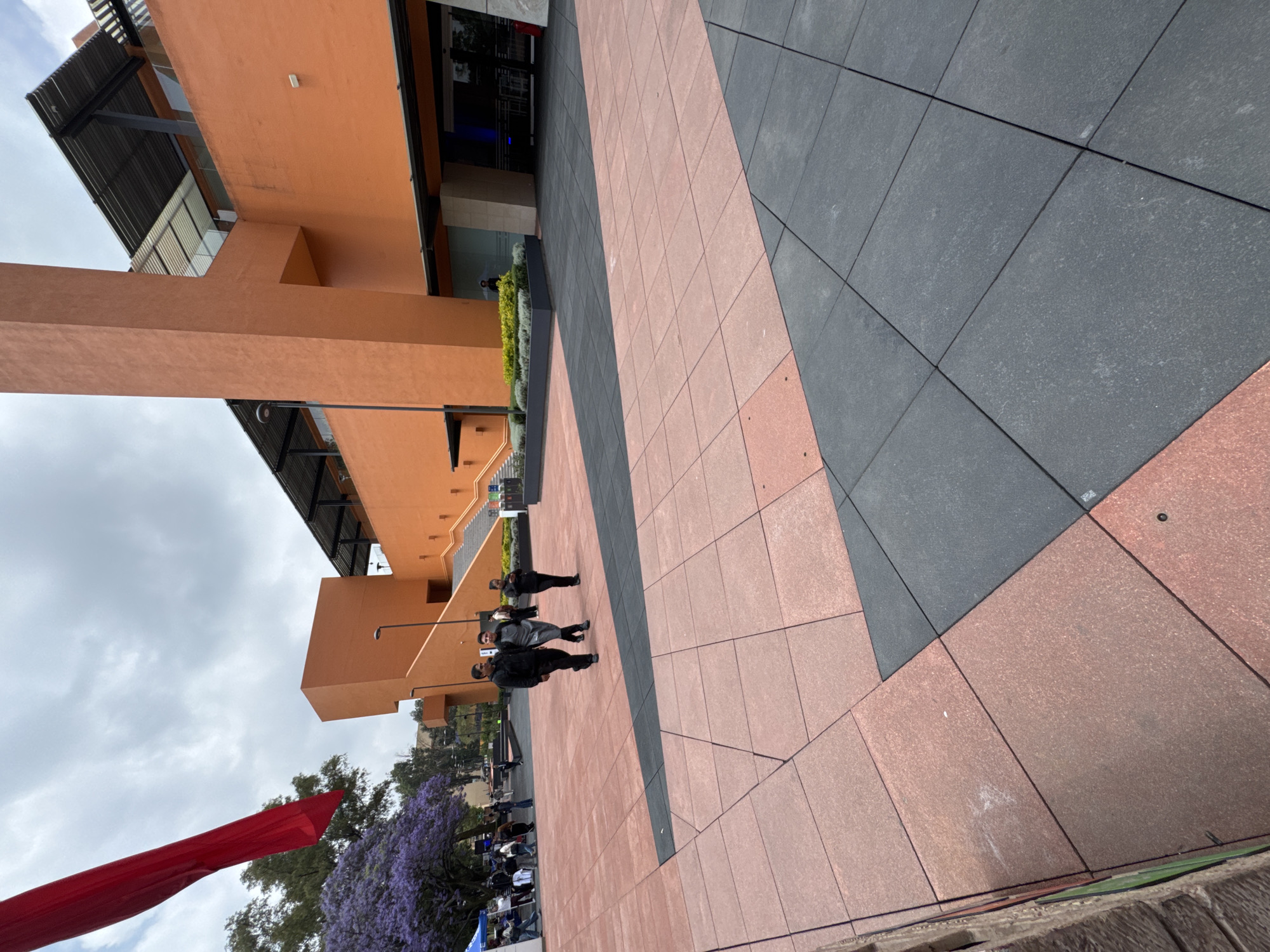 Here is a detailed description of the image:
**General Scene:** The image presents an outdoor urban setting, likely a plaza or pedestrian area in Mexico City, Mexico. The scene is captured from an elevated perspective, looking down at the plaza.
**Architecture:**
* The plaza is surrounded by modern, orange-toned buildings with strong geometric lines.
* The buildings have multiple levels, indicated by the presence of balconies and roof structures.
* There are visible windows and metal awnings, suggesting commercial or institutional purposes.
**Plaza Surface:**
* The plaza is paved with large square tiles of two contrasting colors: pinkish-red and dark gray, arranged in diagonal bands.
**People:**
* Several individuals are walking across the plaza. They are dressed in dark clothing, likely casual attire.
* The people appear to be engaged in normal pedestrian traffic, walking towards or away from the buildings.
**Vegetation:**
* There is a purple blooming Jacaranda tree visible, a characteristic feature of Mexico City.
* Hedges and other greenery are planted around the buildings.
**Other Objects:**
* A portion of a red flag or banner is visible in the lower left corner, potentially indicating an event or decoration.
**Environment:**
* The sky is overcast, with diffuse sunlight illuminating the scene.
* The overall atmosphere suggests a typical day in an urban environment.
**Location Clues:**
* The architectural style, combined with the presence of Jacaranda trees, suggests the location is in Mexico City.
**Actions/Interactions:**
* People are walking around on the plaza.
* The scene captures a moment of daily life in a city square.
In summary, the image depicts a modern plaza with pedestrians in Mexico City, characterized by its distinct architecture, paved surface, and the presence of iconic Jacaranda trees.