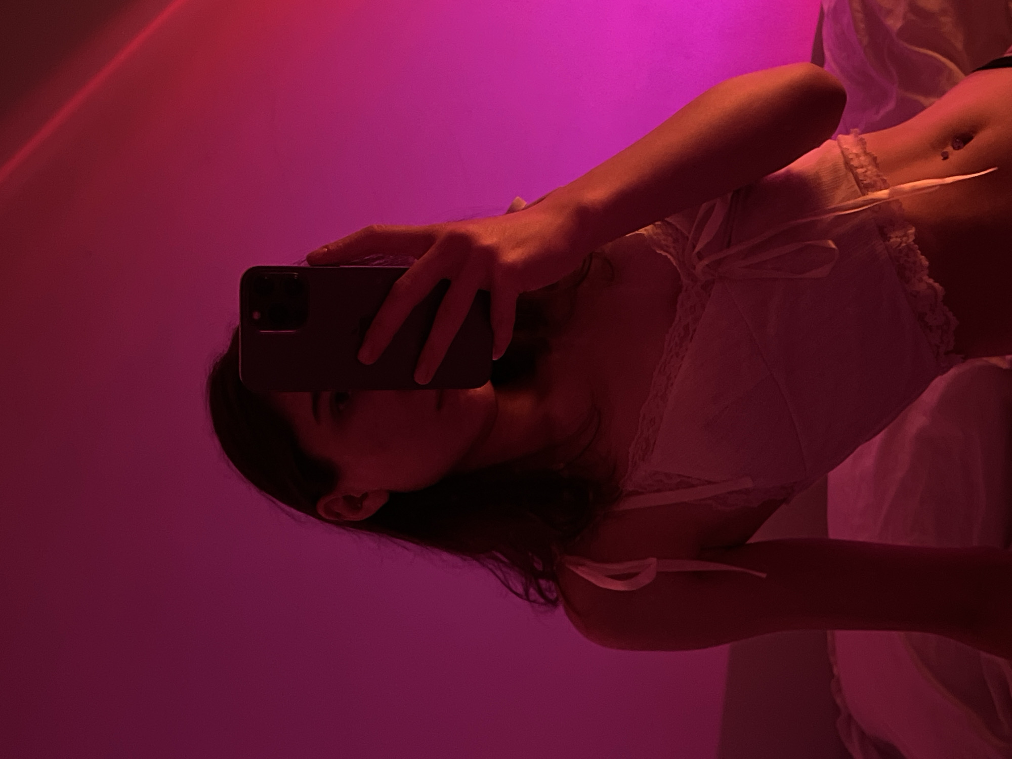 A young woman is taking a selfie in a bedroom. She is holding a smartphone in her right hand and is partially obscuring her face. She is wearing a white camisole. The room is lit with pink and purple light, likely from a sunset lamp. The background is a plain wall. It is not possible to determine the location within Ternopil, Ukraine from the image.