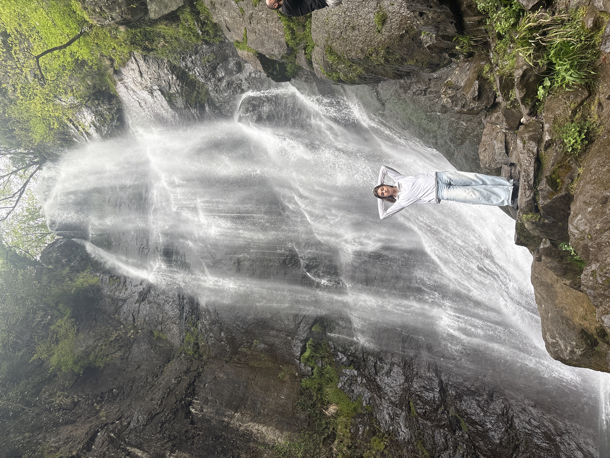 A medium, wide outdoor shot depicts a person posing in front of the Makhuntseti Waterfall in Kveda Makhuntseti, Georgia.
The waterfall cascades down a cliff face. The cliff is characterized by dark, rocky formations and patches of greenery.
A person is lying horizontally on a rocky ledge, appearing to pose in front of the waterfall. They are wearing a light-colored long-sleeved shirt and jeans. Their hands are positioned behind their head.
In the upper part of the frame, obscured by the angle and lighting, are hints of foliage and rocky outcroppings, along with parts of other people.