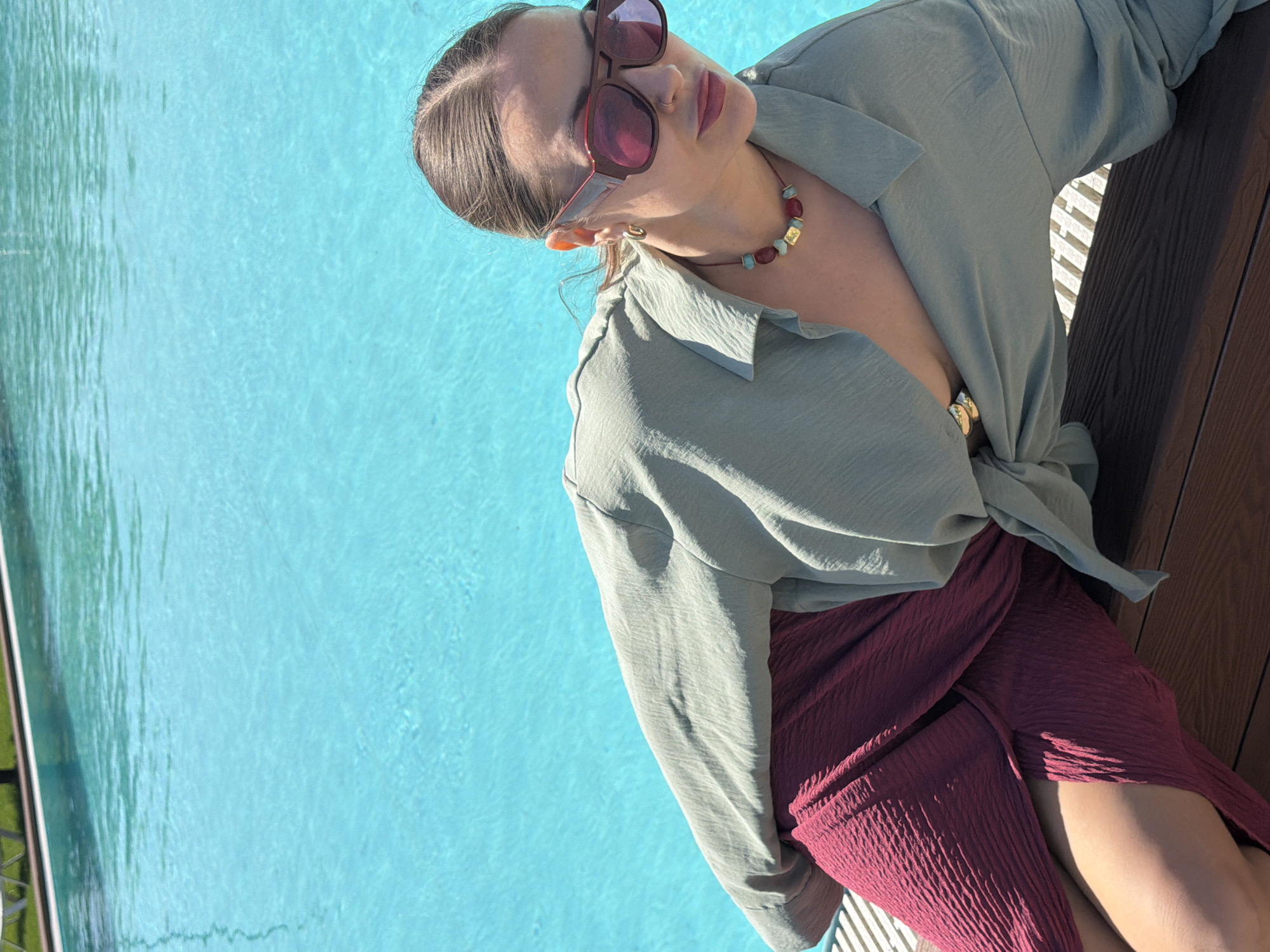The image features a woman posing outdoors by a vibrant blue swimming pool on a bright, sunny day. She is shown from the chest up, leaning against a dark brown wooden surface, with her legs and a portion of her deep red skirt also visible. Her light brown hair is pulled back, and she wears large, reddish-brown framed sunglasses.
A colorful beaded necklace, featuring red, blue/turquoise, and gold-colored stones, is visible around her neck, complemented by a small gold-colored earring on her right ear. She is dressed in a light green, long-sleeved, collared shirt made of a textured, crinkled fabric, which is partially unbuttoned. Her deep red skirt, also of a textured, crinkled material, has a high slit.
The background is dominated by the clear, sun-dappled blue water of the swimming pool, exhibiting ripples and reflections. A metallic fence or railing is faintly visible on the far left edge of the pool. The bright daylight and clear water suggest a sunny weather and a relaxed, leisurely atmosphere, likely at a poolside location such as a resort or private residence. No text is visible in the image.
