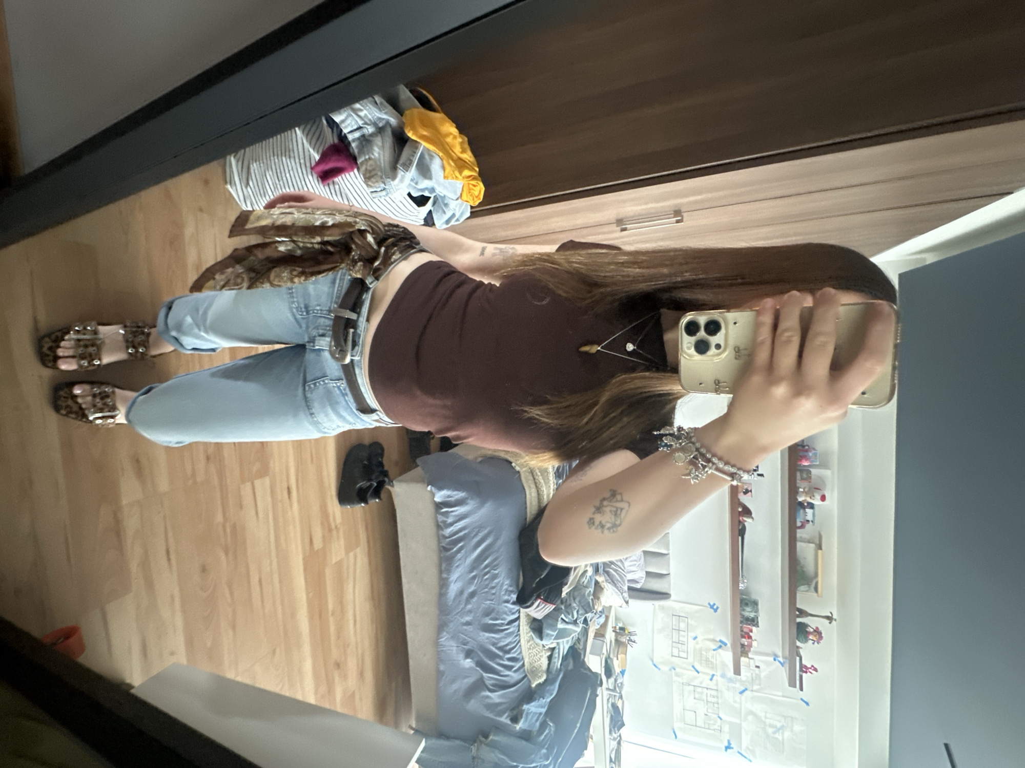 The image shows a person taking a selfie in a bedroom. The person is wearing light blue jeans, a brown top, a brown patterned scarf, and leopard print sandals. They are holding a gold-colored smartphone in their right hand, obscuring part of their face. The background includes a wooden floor, a bed with blue linens, and a wardrobe with open drawers revealing clothes. The room also has a wall with drawings taped to it, shelves with small decorative items, and part of a window. The location is not explicitly mentioned in the image, so the claim that it is in Huixquilucan de Degollado, Mexico, cannot be confirmed from the image alone.