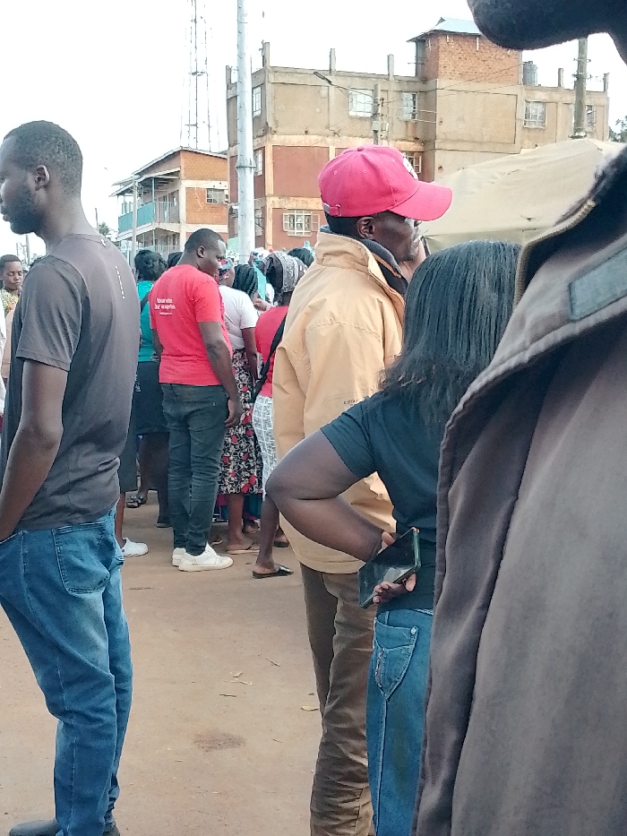 The image captures a group of people standing in a line on a dirt road. The group is mostly women and children. They are wearing casual clothing. The person in the foreground is a man wearing a black shirt and blue jeans. He is looking at the camera. Behind him are people standing in a line. There is a man in a red shirt, a man in a tan jacket and a woman in a black shirt. The woman has her back to the camera and she is holding a smartphone in her right hand. The people are standing on a dirt road. There are buildings in the background. The scene is taking place in the city of Lwandeti, Kenya, which is located at latitude 0.563 and longitude 34.805.