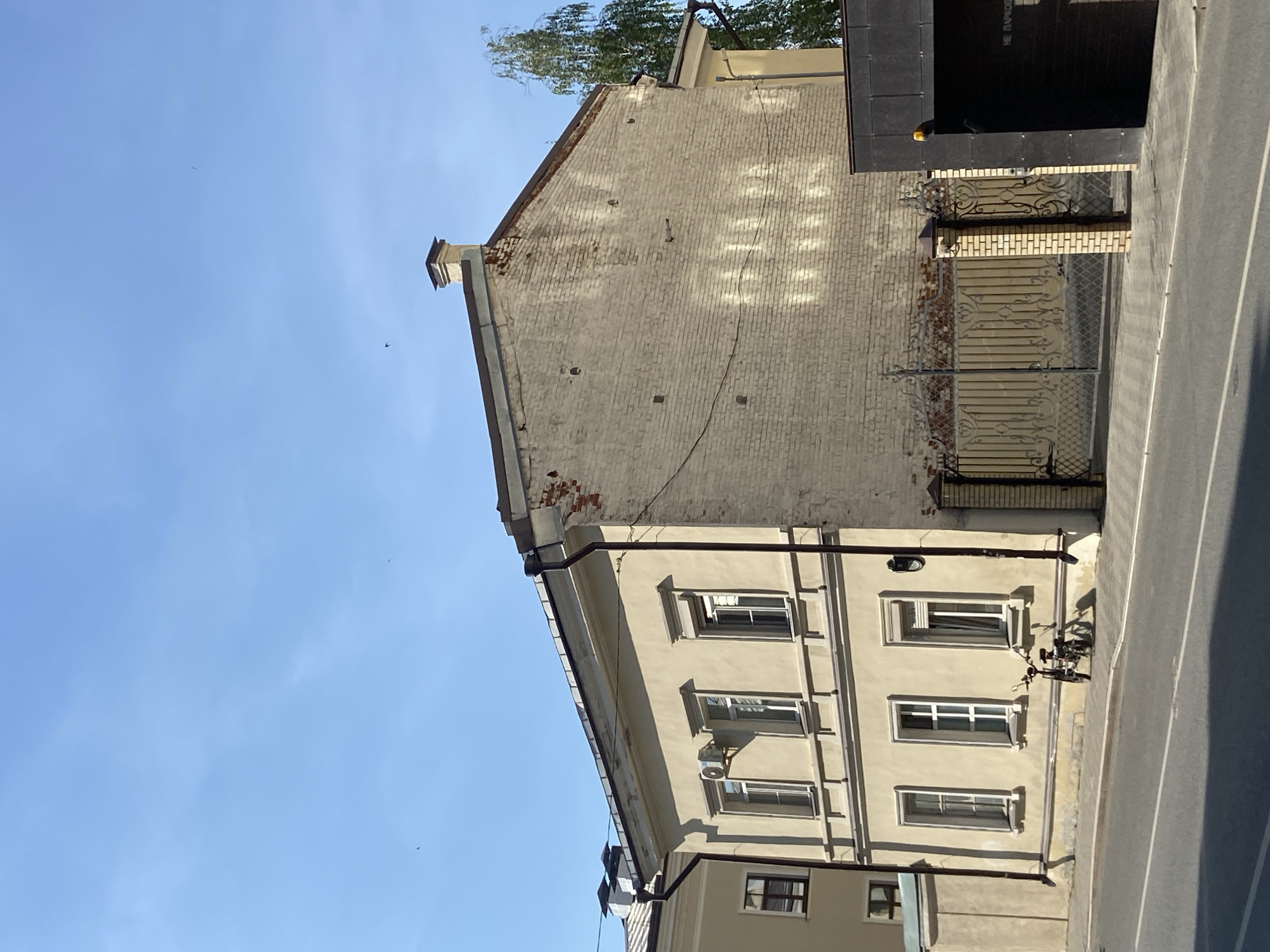The image depicts a two-story building in Kazan, Russia. The building's facade is light beige with symmetrical window placement. An air conditioning unit is visible on the exterior. The upper portion of the building features a taller, aged brick wall with signs of wear. A decorative metal gate adjoins the building, leading to what appears to be a small entrance area. A bicycle is parked adjacent to the building on the sidewalk. The sky is blue and clear.