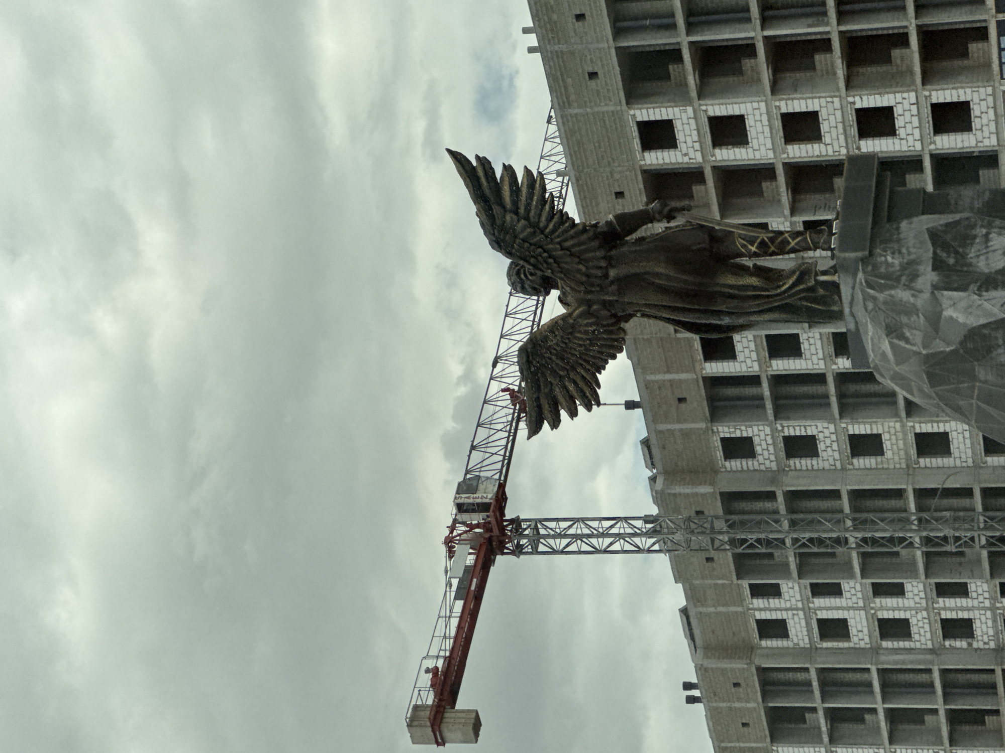 This image shows a statue of an angel with large wings, viewed from the back, standing on a stone pedestal. Behind the statue is a large, unfinished building constructed with light-colored bricks and concrete frames, with many visible window openings. A red and gray construction crane is positioned in front of the building, with its arm extended horizontally. The sky is overcast with gray clouds. The scene is located in the city of Mikhaylovsk, Russia.
