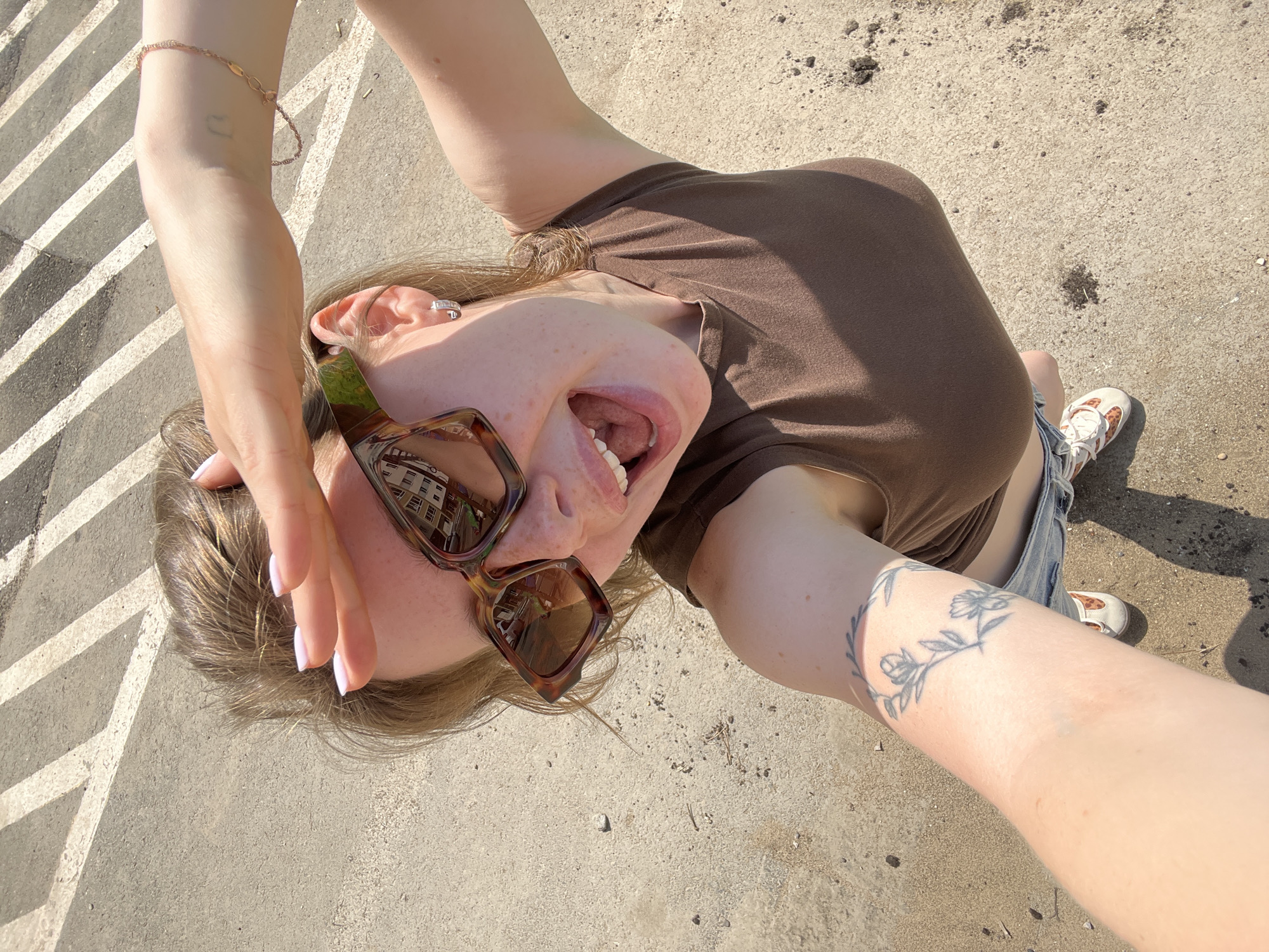 A young woman with blonde hair, wearing sunglasses, a brown tank top, and denim shorts, is taking a selfie outdoors on a sunny day. She is holding her hand up to shield her eyes from the sun, and her mouth is open in a joyful expression. The background shows a cracked concrete surface with white painted lines, possibly a road or parking lot, and some scattered debris. There is a faint tattoo of a floral design on her left forearm. The image does not contain any identifiable landmarks or features that would specifically place it in Chaikovsky, Russia.