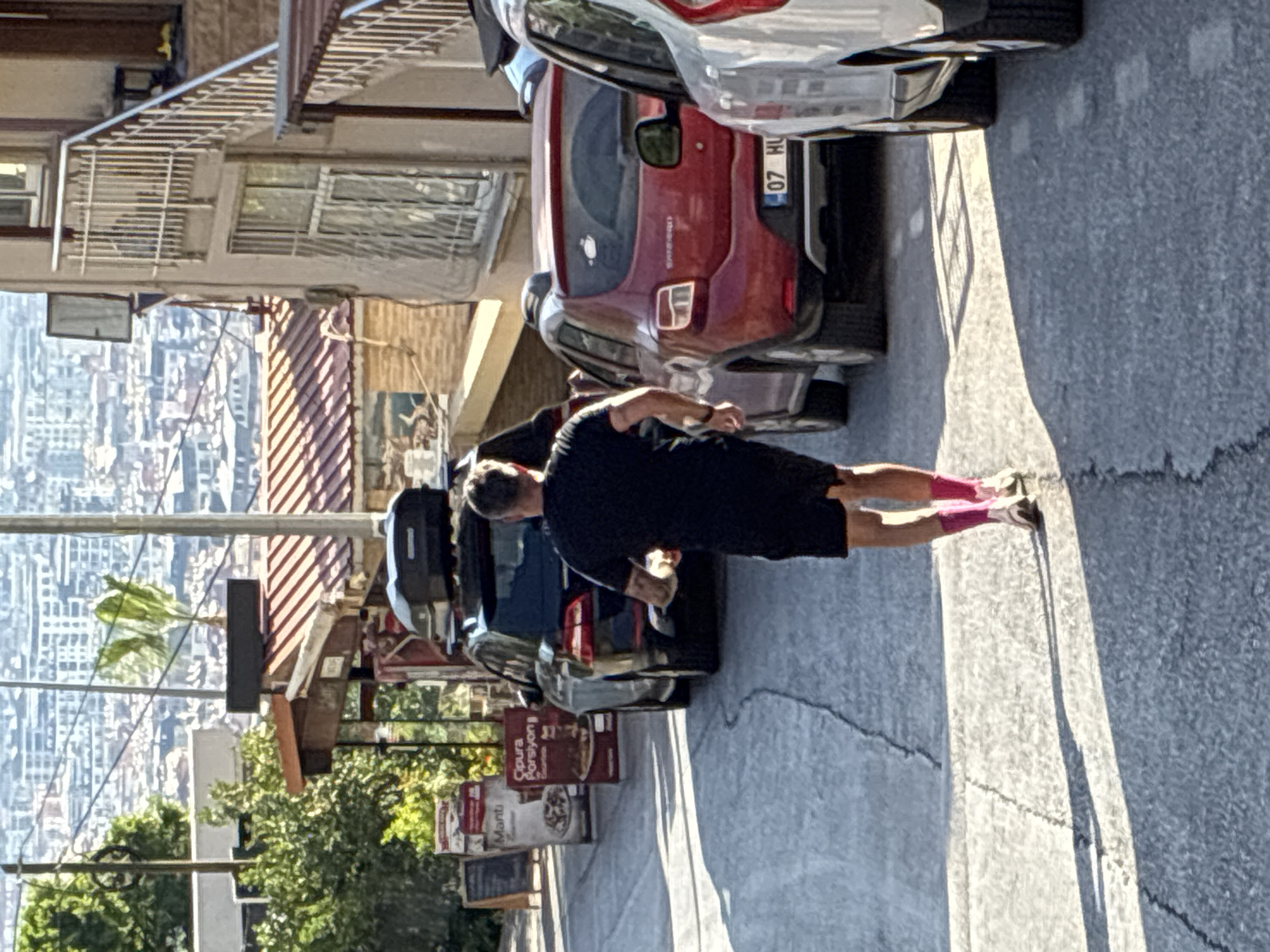 The image shows a man wearing a black t-shirt, black shorts, and pink socks, running away from the camera down a paved street in Alanya, Turkey. He is holding a clear plastic bottle in his right hand. The street is lined with buildings on the right and trees and signs on the left. Several cars are parked on the street, including a black SUV with a roof rack, a red hatchback, and a white car. In the background, the city of Alanya is visible with its buildings and a distant view of the sea. The lighting suggests it is a sunny day.