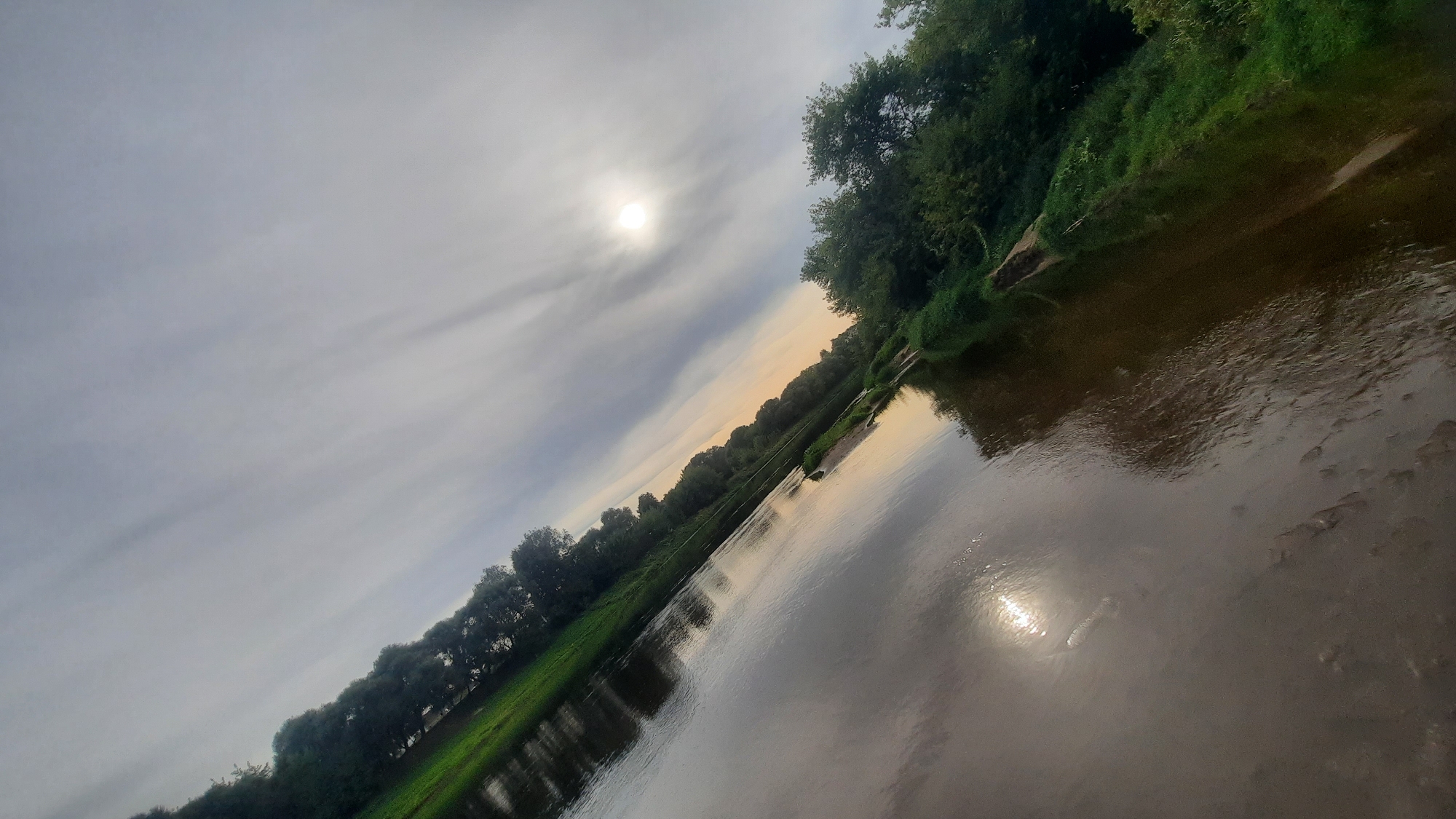 The image depicts a river flowing through a green, tree-lined landscape. The sky is cloudy with the sun visible, casting reflections on the water. The camera angle is tilted, giving a dynamic perspective of the scene. The riverbank on the left is lush with grass and trees, while the right bank features denser foliage. The water appears calm, with ripples on the surface. The overall impression is one of natural beauty and tranquility. The image is a photograph taken from an elevated position, possibly a bridge or a nearby hill.