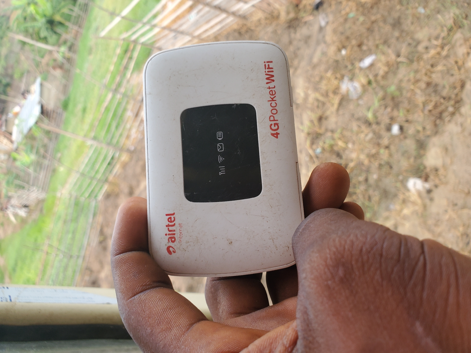 This image depicts a close-up view of a dark-skinned hand holding a white 4G Pocket WiFi device, set in an outdoor environment in Winters, United States of America. The device prominently features the 'airtel' logo with 'Internet' below it in red text on its upper left front surface. Its central black LCD screen displays active full signal strength, a Wi-Fi symbol, a message icon, and a full battery indicator. Below the screen, "4G Pocket WiFi" is printed in red. The device itself shows signs of wear, including multiple scratches and surface dirt. The hand gripping the device is positioned in the lower left and center of the frame, showing distinct finger creases and skin texture. The background is out of focus, revealing a natural outdoor setting with dry, light brown ground interspersed with sparse vegetation and small pieces of debris directly behind the device. Further back, a rustic wooden fence or barrier extends across the middle ground, leading to an area of green vegetation and a light-colored structure with a metallic roof. A vertical tan-colored pole or structural element is partially visible on the far left edge of the image.