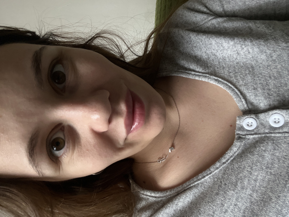 An adult female is depicted in a close-up, head-and-shoulders portrait, taken indoors in Leninakan, Russia. She has brown eyes, a light complexion, and dark brown hair that frames her face. Her lips appear full with a glossy sheen, and her expression suggests a slight, closed-mouth smile. She wears a grey, heathered long-sleeved top with a rounded neckline, featuring two visible white circular buttons on a vertical placket at the chest. A thin silver-toned chain necklace with a small, clear faceted stone pendant is positioned on her lower neck, with its clasp mechanism visible to the left. A small silver-toned hoop earring is present in her left earlobe. The background is out of focus, consisting of a plain, light-colored surface on the left and a green, textured fabric object on the right. Lighting appears soft and frontal.