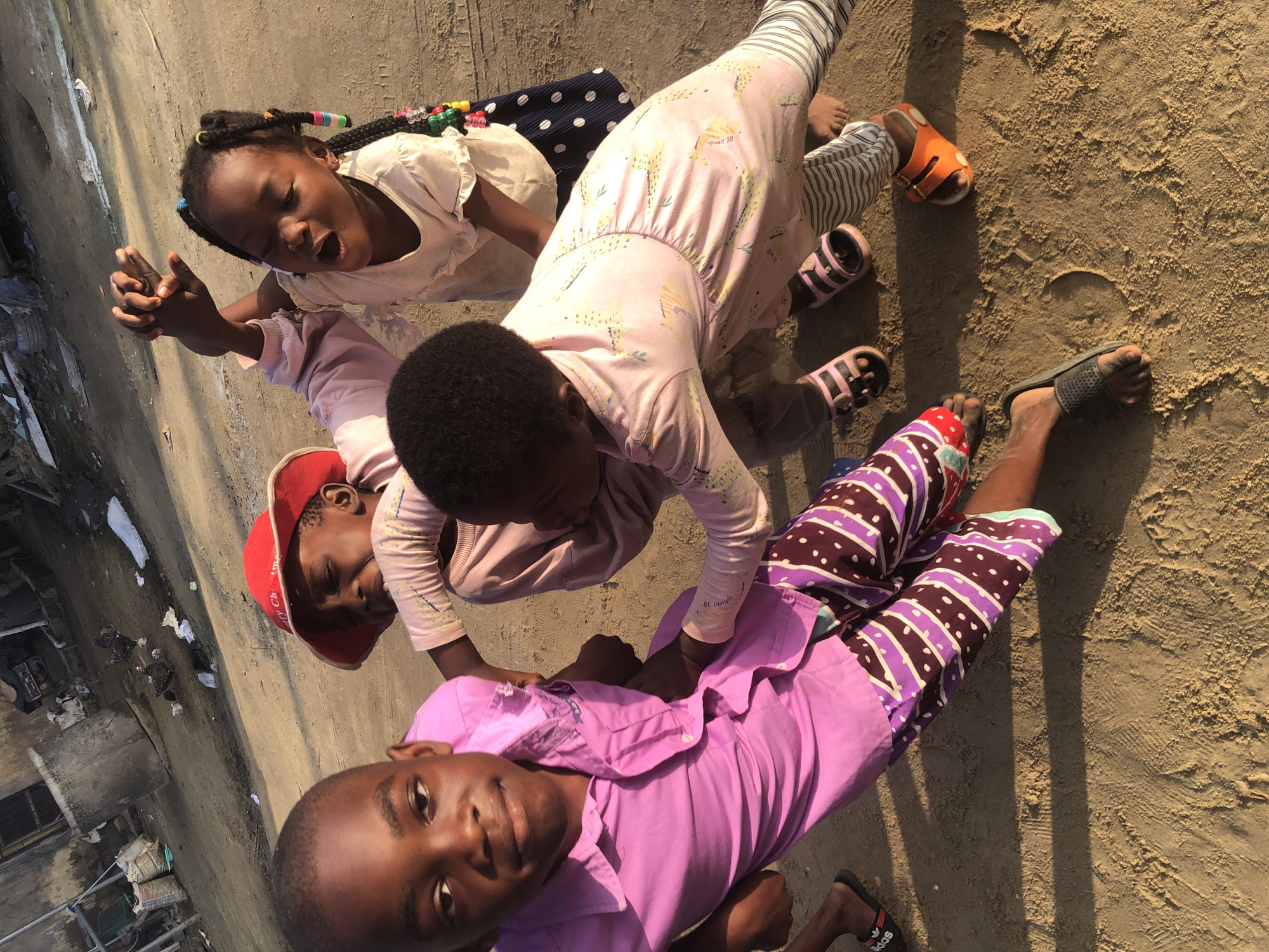 The image captures a group of four young children interacting on a sandy ground, possibly in Ado, Nigeria. In the foreground, a boy in a vibrant pink collared shirt and patterned trousers is looking towards the camera with a slight smile. His left hand is extended, and his right arm is being held by another child.
To the left and slightly behind him, a child wearing a red hat and a light-colored shirt is looking down and appears to be crying or distressed. This child is also being touched by another child.
In the center and to the right, two girls are engaged with each other and the other children. One girl, wearing a light-colored dress, has her arms raised and is looking upwards with her mouth open. The other girl, wearing a pink patterned outfit, is leaning forward, her arms around another child. Both girls have braids adorned with colorful beads. The ground beneath them is a mix of sand and dirt, with footprints and tire tracks visible. In the background, there are indications of a somewhat dilapidated urban or semi-urban environment, with concrete structures, debris, and what appear to be some animals, possibly chickens and a cow, suggesting a rural or peri-urban setting. The lighting indicates it is daytime, likely late afternoon given the warm glow.