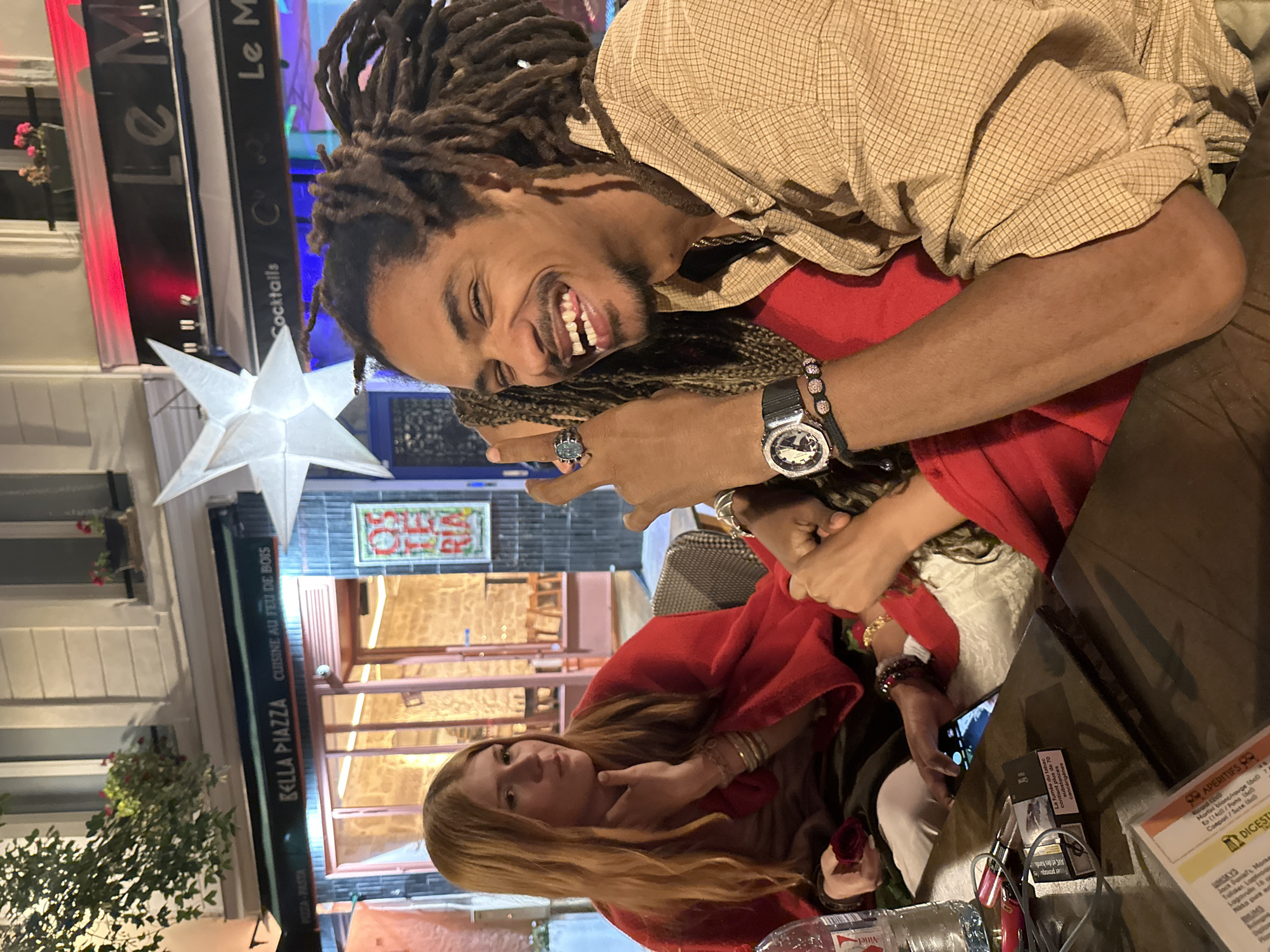 An outdoor evening scene captures two individuals, a man and a woman, seated at a table, likely at a restaurant or cafe in Paris, France. The time of day is clearly night, indicated by the use of artificial lighting and the dark background.
The man, with dreadlocks and a beard, wears a light brown checkered shirt over a red top. He is smiling broadly towards the camera, his arm around the woman. He sports a silver-toned ring on his left hand and a watch and a beaded bracelet on his left wrist.
The woman, with long blonde hair, is dressed in a red shawl or cardigan. She looks towards the camera with a neutral, contemplative expression, her hand near her chin. She wears bracelets on her wrist and holds a smartphone.
On the dark tabletop, a bottle of "Vittel" water is visible, alongside glasses, a cigarette packet with French health warnings ("La fumée du tabac contient plus de 70 substances cancérigènes"), and a menu with sections like "APÉRITIFS" and "DIGESTIFS".
The background features illuminated buildings and restaurant signs with French text. A prominent white star-shaped light fixture hangs nearby. Visible signs include "Le M" (appearing twice), "Cocktails", and "BELLA PIAZZA CUISINE AU FEU DE BOIS", which also mentions "PIZZA-PASTA". Another tiled sign with "RIA" is partially visible. The overall atmosphere suggests a casual, social dining experience.
