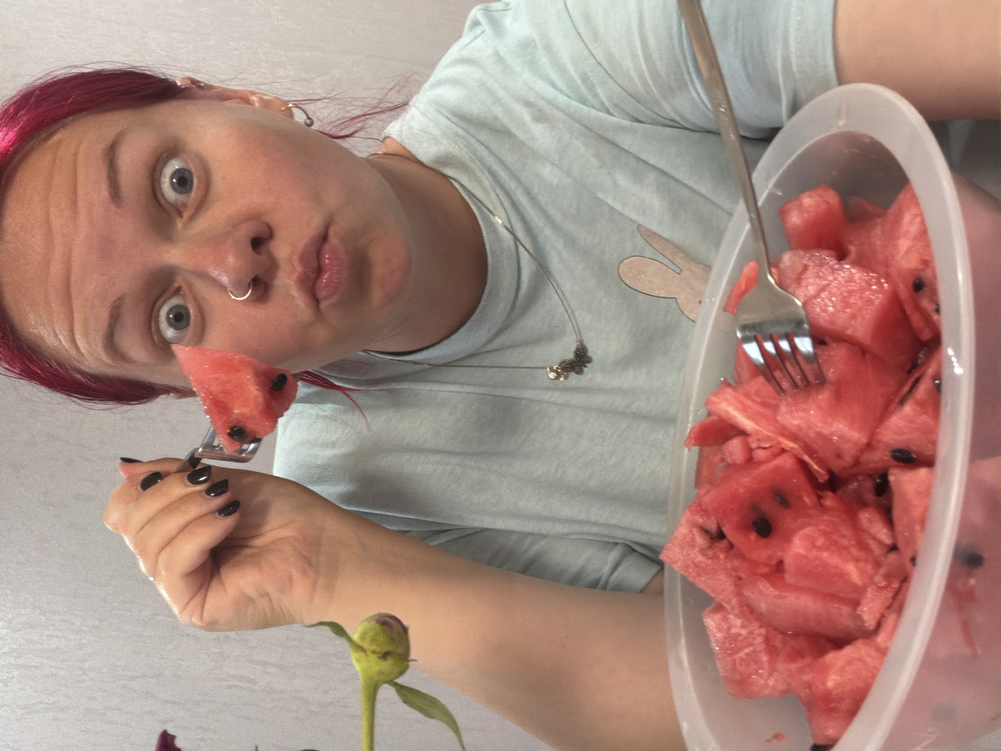 A Caucasian female with bright pink hair, a nose ring, and earrings is looking directly at the camera with wide eyes and a puckered mouth, holding a piece of watermelon on a fork. She is wearing a light blue t-shirt with a graphic of a bunny. In front of her is a clear plastic bowl overflowing with cut-up watermelon pieces. A fork is resting in the bowl. To the left of the woman, a small green plant with a bud is visible. The background appears to be a light-colored, textured wall. The image is a close-up shot, focusing on the woman and the watermelon. The location is not specified in the image.