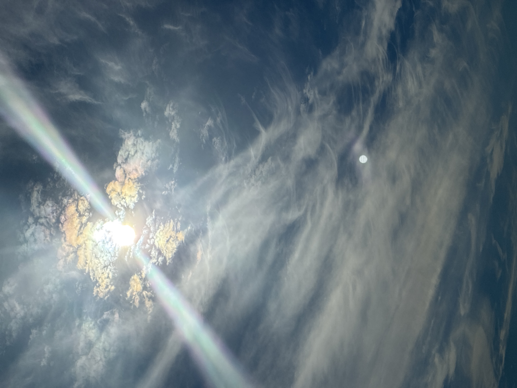 A low-angle, vertical shot captures a dramatic sky. The sun is a bright, blown-out white orb at the top left, partially obscured by wispy, iridescent clouds that glow with yellow, orange, and pink hues. A broad, diagonal band of light, exhibiting a rainbow effect, streaks across the upper portion of the frame, originating from the sun and extending towards the bottom right. The sky itself is a deep blue, filled with delicate, feathery clouds that form swirling patterns. In the lower right quadrant, a small, distinct circular object with a patterned surface is visible, appearing to be either a distant celestial body or an artificial light source. The overall impression is one of atmospheric grandeur and celestial phenomena. The location is unknown.