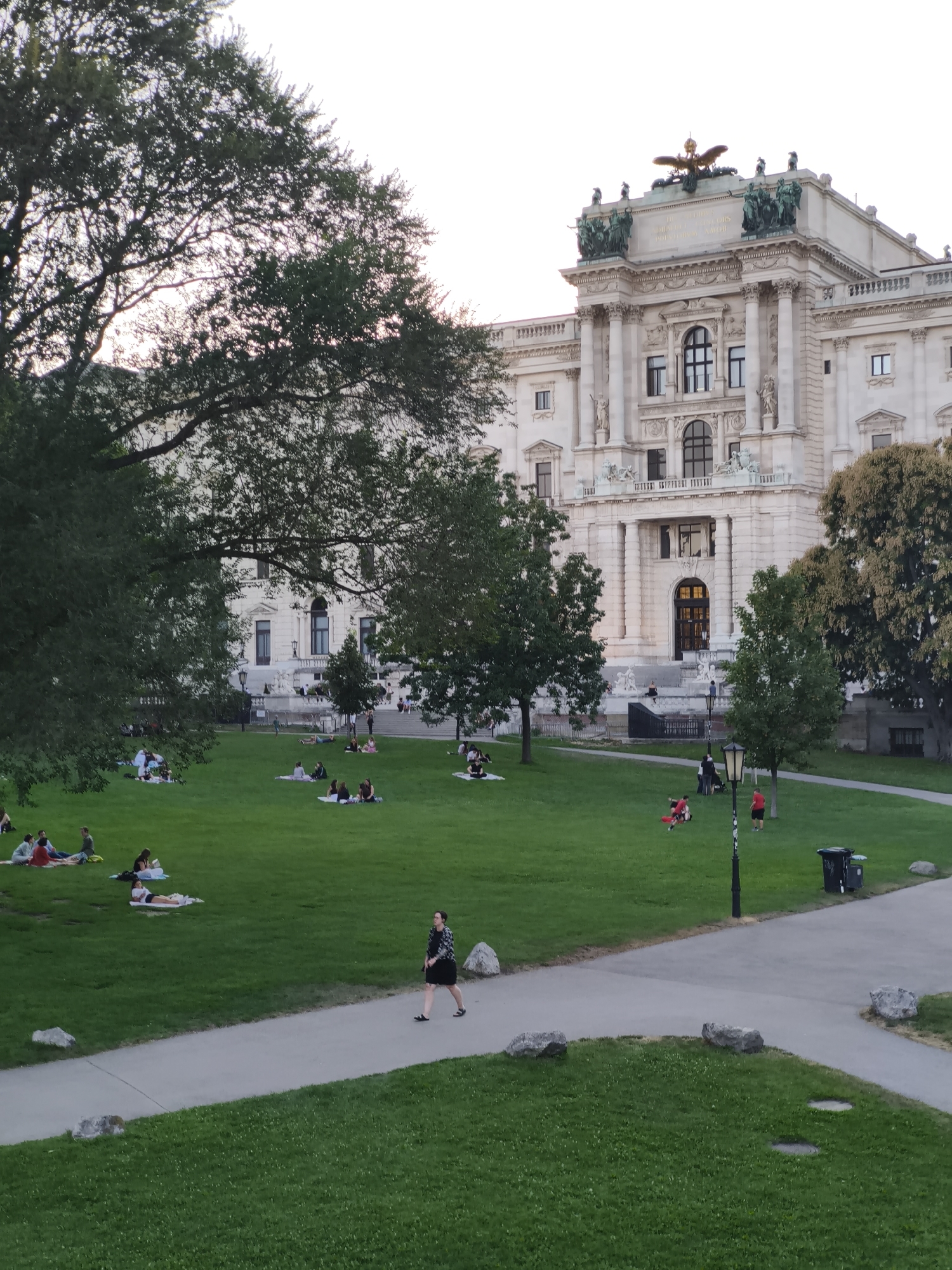 The image depicts a bright, outdoor scene in Vienna, Austria, likely in the late afternoon or early evening, with a large, ornate building in the background. The building is made of light-colored stone and features classical architectural elements such as columns, intricate carvings, and statues. A prominent golden eagle statue sits atop the roof.
In the foreground and midground, a spacious green lawn is dotted with numerous people engaged in various leisurely activities. Many are sitting or lying on blankets, appearing to be picnicking or relaxing. A paved pathway winds through the lawn, and a few individuals are walking or standing along it. One person in the midground is walking on the path, wearing a dark patterned top and a black skirt. Another person in the background is seated on the path, facing away from the camera.
Several trees are scattered across the scene, providing shade and framing the view of the building. The overall atmosphere suggests a pleasant day in a public park or square, with people enjoying the open space and the impressive architecture. A trash bin is visible on the right side of the pathway.