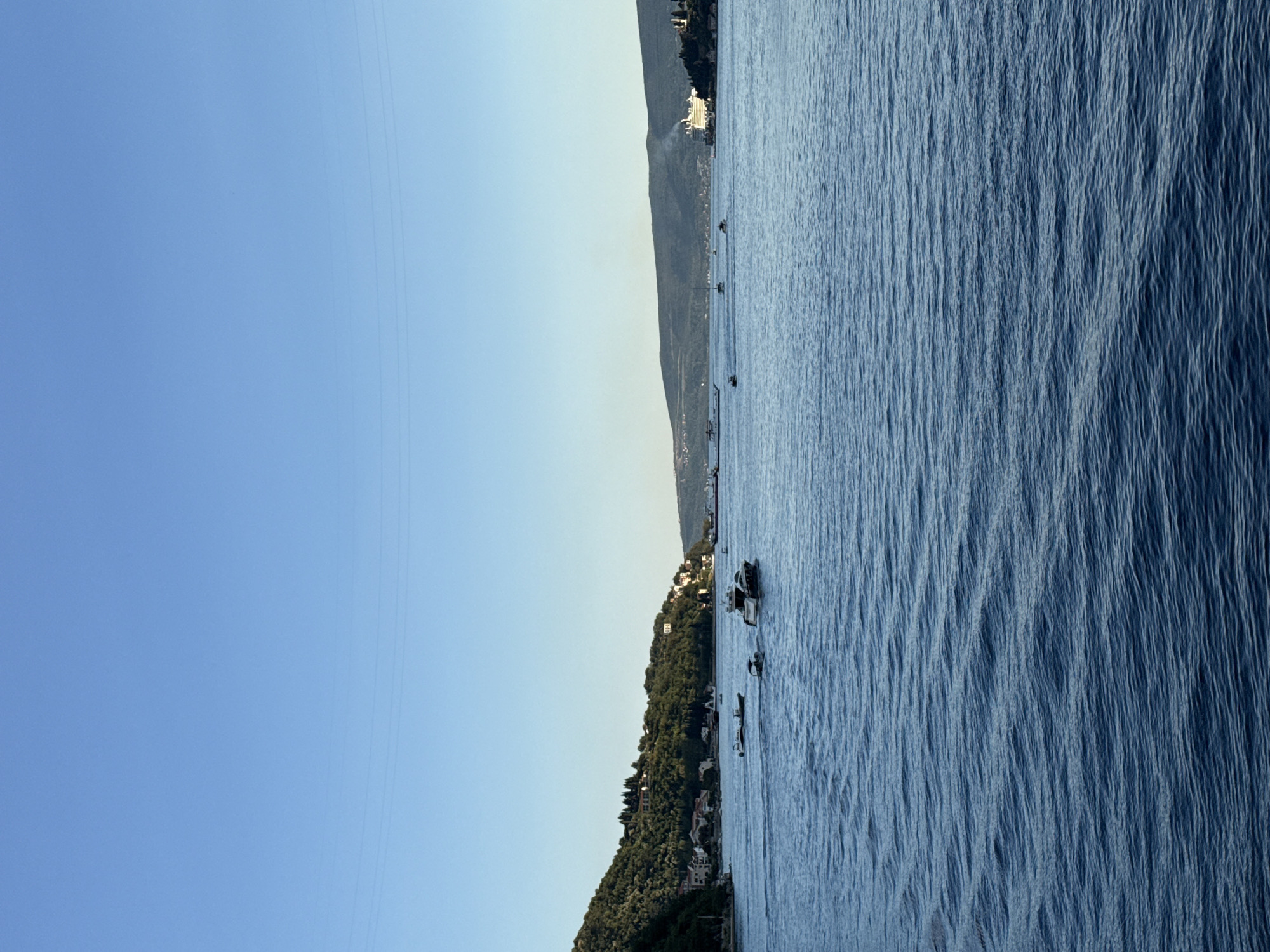 A wide-angle, eye-level shot captures a serene bay in Đurići, Montenegro, under a clear, pale blue sky. The foreground is dominated by the tranquil, deep blue water of the bay, rippled by a gentle breeze. Several boats of varying sizes are scattered across the water. In the middle ground, on the left side of the frame, a densely forested green hillside rises, dotted with scattered houses with red tile roofs, indicating a coastal settlement. On the right side of the bay, a long, low mountain range stretches across the horizon. A large white cruise ship is visible in the far distance on the right, emitting a plume of white smoke. The overall impression is one of calm and natural beauty, with human habitation integrated into the landscape.