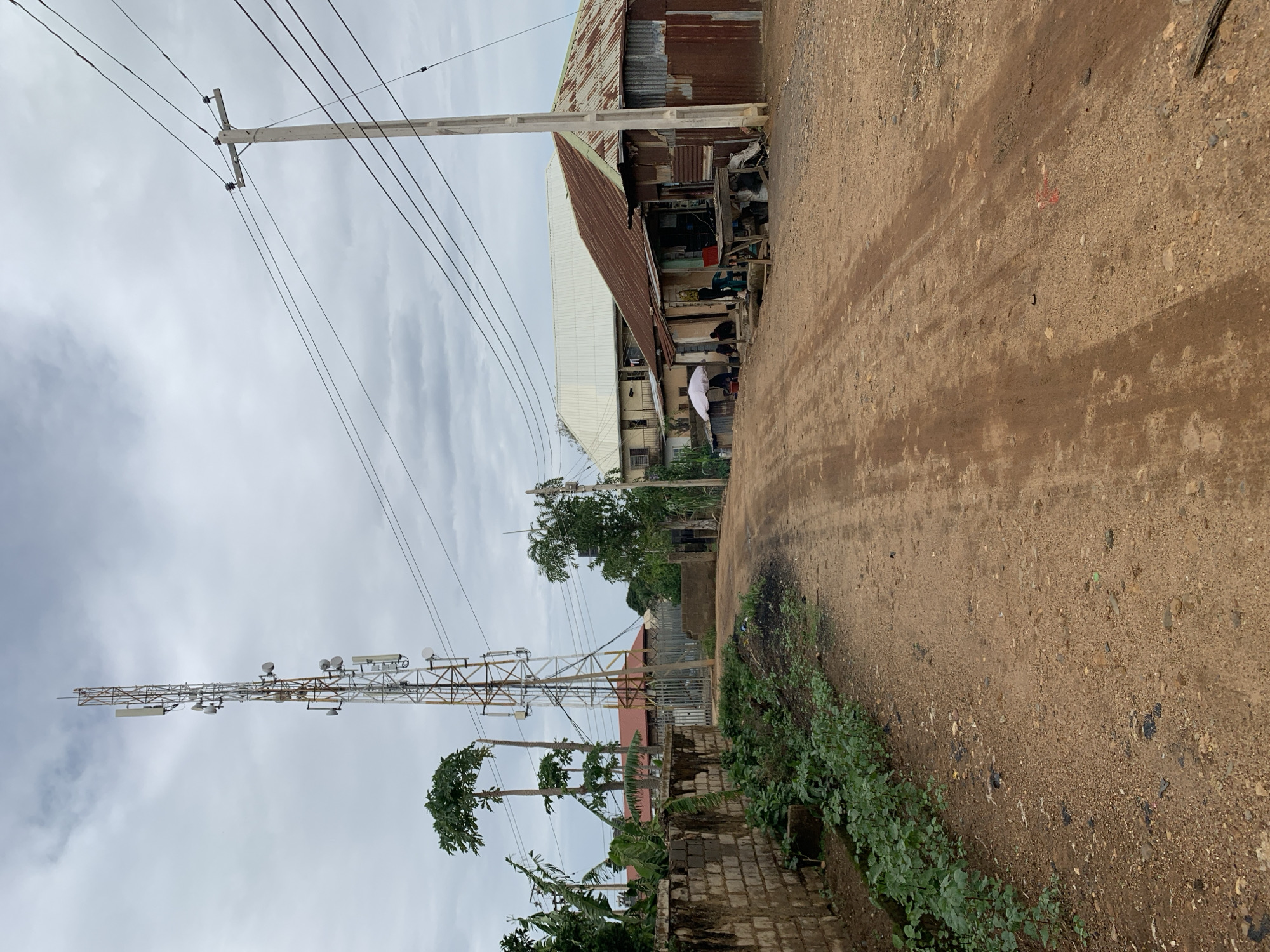 This image captures a street scene in Rafin Par, Nigeria, under an overcast sky. The foreground is dominated by an unpaved, reddish-brown dirt road exhibiting distinct tire tracks and scattered small stones, receding into the distance with a slight curve to the right.
On the left side, a low, unplastered brick wall is partially obscured by green leafy plants growing along its base. Further along this side, a tall, metallic lattice communications tower, featuring multiple antennae and dishes, stands prominently; visible rust indicates its age. A metal gate or fence is situated near the tower. Several tall, slender trees, possibly papaya, are present, and red-roofed buildings are discernible in the background.
On the right side, a concrete utility pole with multiple overhead power lines extends diagonally across the upper portion of the frame. Adjacent to the road, a single-story structure constructed from a combination of rusted and galvanized corrugated metal sheets serves as a roadside shop, displaying its contents through an open front. Several individuals are present in front of and within this structure: one person is seated under a white umbrella, another stands near the entrance, and at least two additional people are visible inside or close to the shop. Behind this, a larger, multi-story building with a light-colored corrugated metal roof and balconies is visible. The scene depicts typical daily life along an unpaved local thoroughfare.