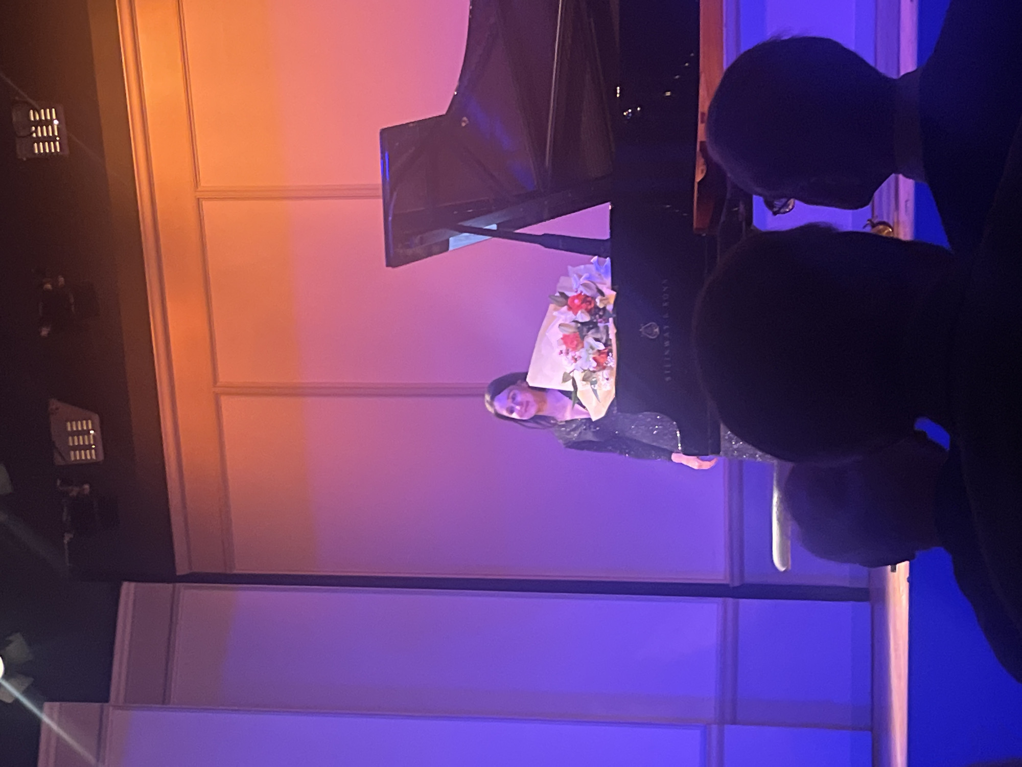 A vertical indoor shot shows a female performer holding flowers next to a Steinway & Sons piano, with the partial silhouettes of audience members in the foreground. The background features paneled walls lit with orange and purple light. The location is potentially a concert hall in Berlin, Germany, given the make of the piano.