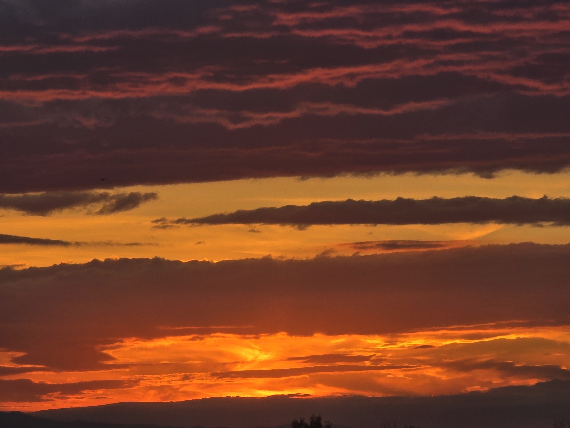 The image depicts a vibrant sunset with layered clouds. The top layer of clouds appears in shades of deep purple and red, arranged in horizontal bands. Below, a broad band of orange is punctuated by grey cloud formations. The lower portion of the image showcases clouds in vivid reds and oranges, with the light emanating from the setting sun partially obscured by these clouds. In the far distance, a dark horizon is visible, topped with minimal vegetation. The location is unknown.