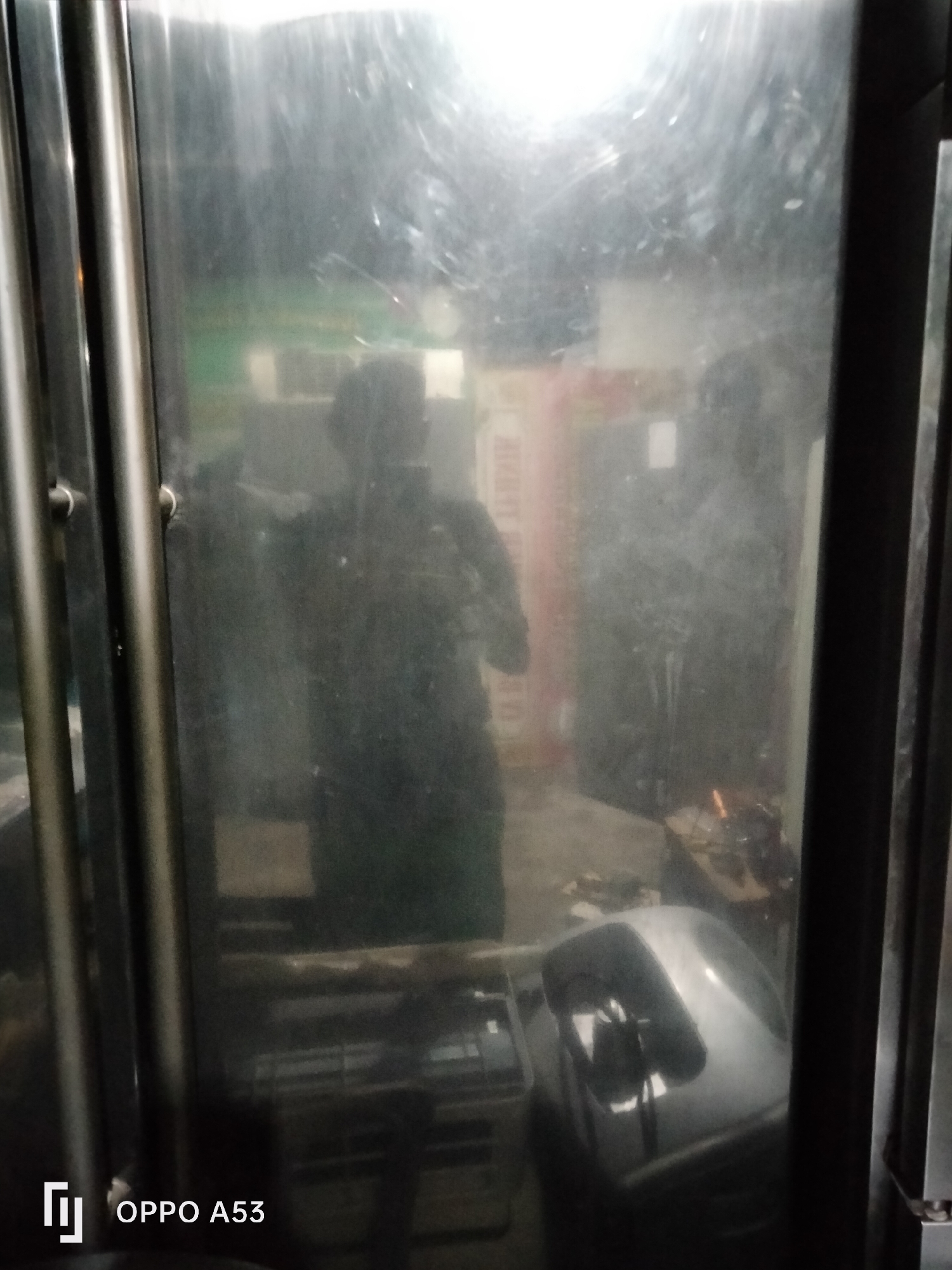 The image is a low-light, reflective shot taken from within a metallic enclosure, possibly a lift or elevator cabin. The reflective surface of the interior walls obscures much of the detail, creating a hazy and distorted view of the surroundings.
In the foreground, on the left, are vertical, cylindrical metallic poles, likely part of the cabin's structure. The right side of the image is dominated by a large, reflective surface.
Reflected within this surface, a person is visible as a dark silhouette. They appear to be standing and looking outwards. Beyond the person's reflection, the interior of what seems to be an industrial or commercial space is vaguely discernible. There are shapes that could be shelves, equipment, or furniture. A light source, possibly overhead, casts a bright glare near the top of the reflective surface, washing out details.
In the lower right portion of the image, a curved, metallic object with a reflective surface is visible, possibly part of a vehicle or machinery. There is also a suggestion of a workbench or table with some items on it in the background.
The overall impression is one of a dimly lit, functional space, with the reflections dominating the visual information. The image is marked with the "OPPO A53" watermark in the bottom left corner, indicating the device used for capture.
Without additional context or clearer visual cues, it is impossible to determine the specific location within Denpasar, Indonesia. The scene is generic enough to be found in various urban industrial or commercial settings.
