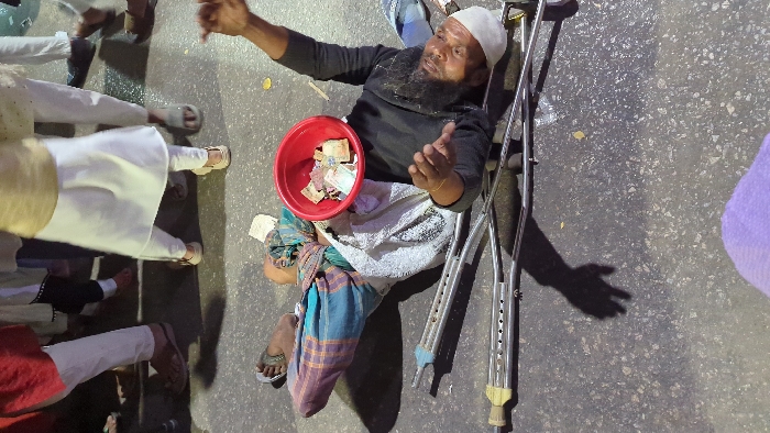 An elderly man with a beard and a white cap is lying on the ground next to a pair of crutches. He's wearing a black top and has a red bowl filled with money resting on his legs. He appears to be gesturing with his hands, possibly begging. Several pairs of legs and feet of people wearing traditional clothing are visible in the background. The scene is set on a paved surface.