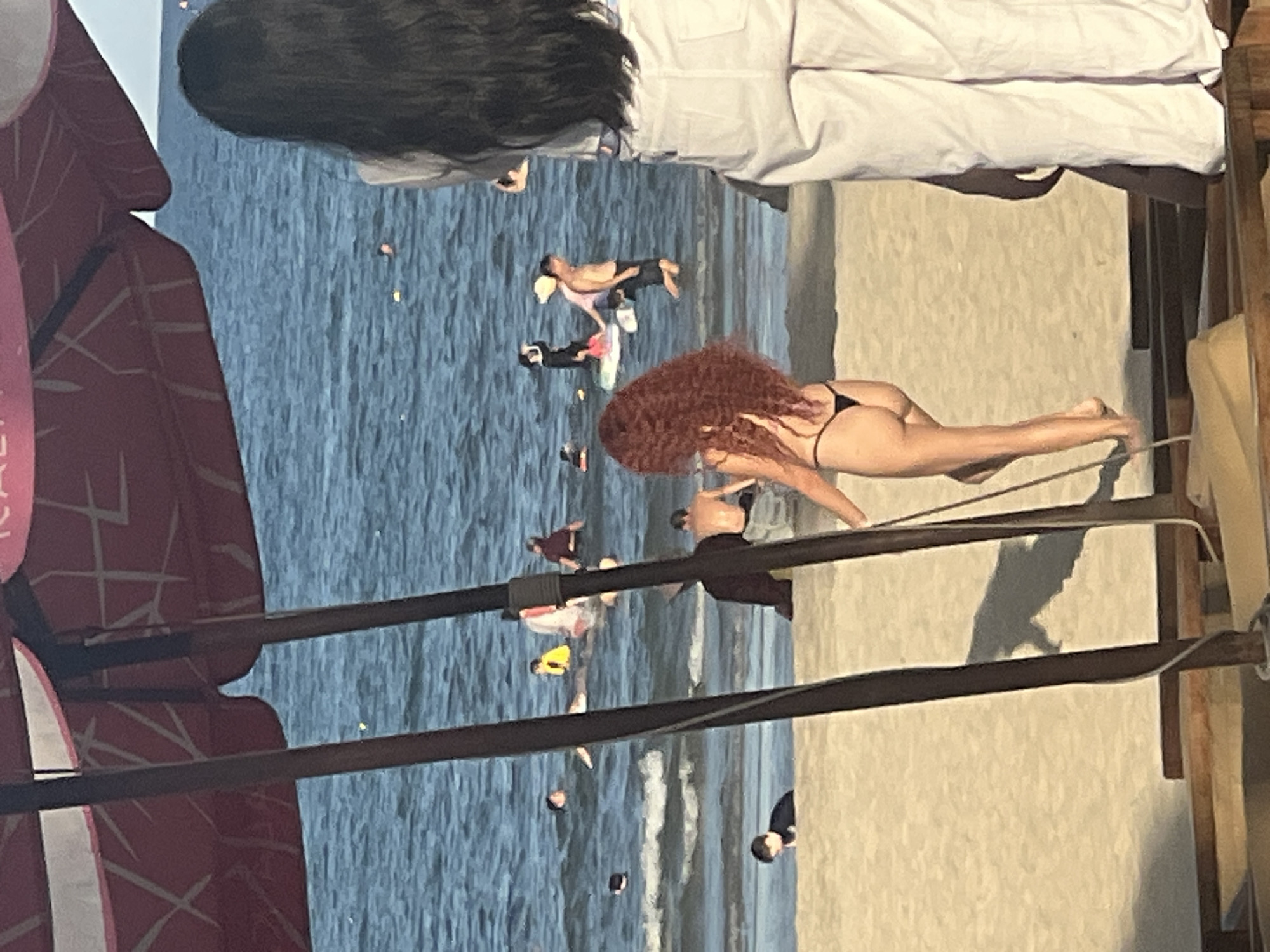 The image depicts a beach scene in Da Nang, Vietnam, with the ocean in the background and a sandy beach in the foreground. Several people are visible in the water, some swimming and others standing. A woman with long, dark hair is standing on the right side of the frame, facing away from the camera. In the middle of the frame, a woman with curly red hair is walking on the beach, wearing a black bikini. Two wooden poles, likely part of a beach umbrella or structure, are in the foreground. The water is blue with some waves. The sky is not visible.