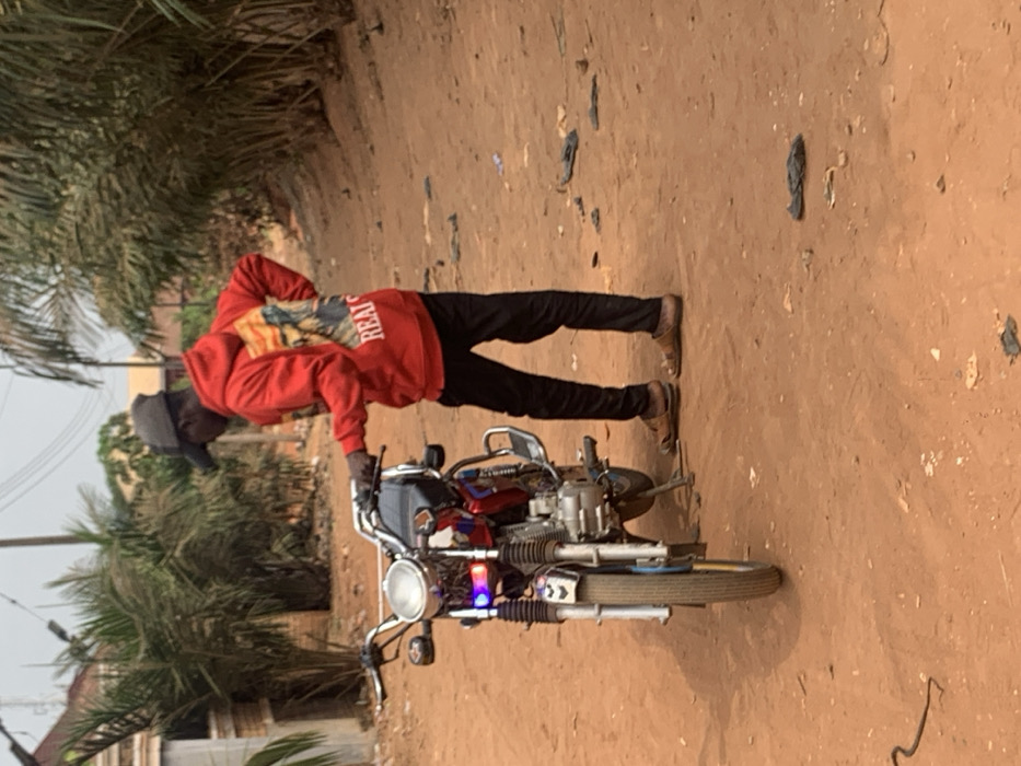 An outdoor scene in Otuocha, Nigeria, features a male individual standing beside a stationary red and silver motorcycle on an unpaved, reddish-brown dirt road. The individual is clad in a red hooded sweatshirt with a graphic on the back depicting a figure and the visible text "REALS," black trousers, brown open-toed sandals, and a dark-colored baseball cap. His left hand rests on the motorcycle's handlebars, while his right hand is placed on his hip. The motorcycle, oriented towards the viewer, has a prominent round headlight, with blue and purple accent lights positioned below it. Its kickstand is deployed. Dense green foliage, primarily palm trees, borders the dirt road, particularly on the right side of the frame. A light-colored building is partially visible on the far left. Scattered dark debris, possibly plastic, is visible on the ground. The ambient lighting suggests clear daytime conditions.