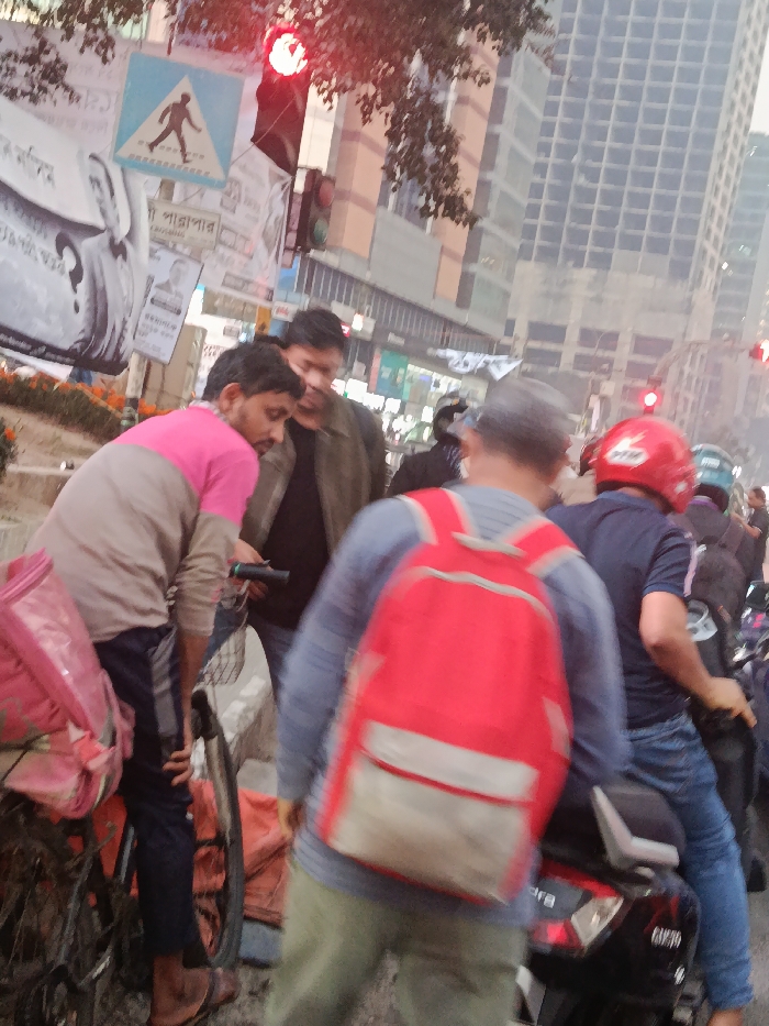 The image captures a detailed street scene in Dhaka, Bangladesh, during daylight hours, likely at a traffic intersection where vehicles are stopped. A red traffic light is visibly illuminated in the upper left, indicating a stop signal.
In the immediate foreground-left, a male individual with a beard, wearing a pink and beige long-sleeved shirt and dark pants, stands beside a black bicycle. He is observed looking downward, holding a dark, cylindrical object in his hands. A pink plastic bag, possibly a delivery carrier, is attached to the rear rack of his bicycle. Adjacent to him, a second male, dressed in a dark jacket over a dark shirt, also directs his gaze downwards, appearing to interact or observe the same point as the first individual.
In the foreground-right, a third male is seen from the back, wearing a light blue long-sleeved shirt, olive-colored pants, and a prominent red backpack with white accents. He is standing in close proximity to a stationary black scooter, identified by a visible "Mahindra" logo on its rear side. Directly behind this individual, a fourth male is seated on the black scooter, wearing a dark blue short-sleeved shirt, blue jeans, and a red motorcycle helmet. His right hand is positioned on the handlebar.
Further in the mid-ground and background, additional male individuals on motorcycles are visible, queued in traffic. One notable rider wears a green helmet.
Above the scene on the left, a blue square traffic sign with a white border and a black pedestrian symbol is affixed to a pole. This pole is also covered with multiple white rectangular posters featuring Bengali script and portrait photographs. A low concrete median strip, adorned with orange flowering plants, runs along the left side of the street. In the background, several modern, multi-story buildings with grid-patterned facades dominate the urban skyline.