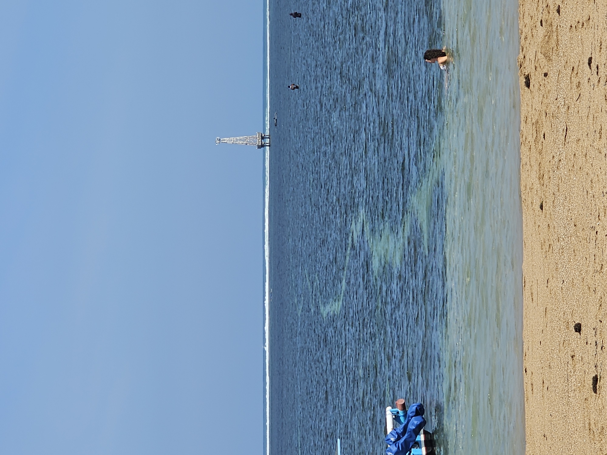 A woman is swimming in the ocean near a beach in Denpasar, Indonesia. The water is clear and blue, with a sandy bottom visible near the shore. A boat is docked on the left side of the image, partially covered in a blue tarp. In the distance, a tall, white tower stands in the ocean, possibly a lighthouse or a research platform. Several other people are in the water, some swimming and others standing. The sky is clear and blue, indicating a sunny day. The waves are small and gentle, breaking on a reef in the distance. The sand on the beach is light brown and speckled with small pebbles and debris.