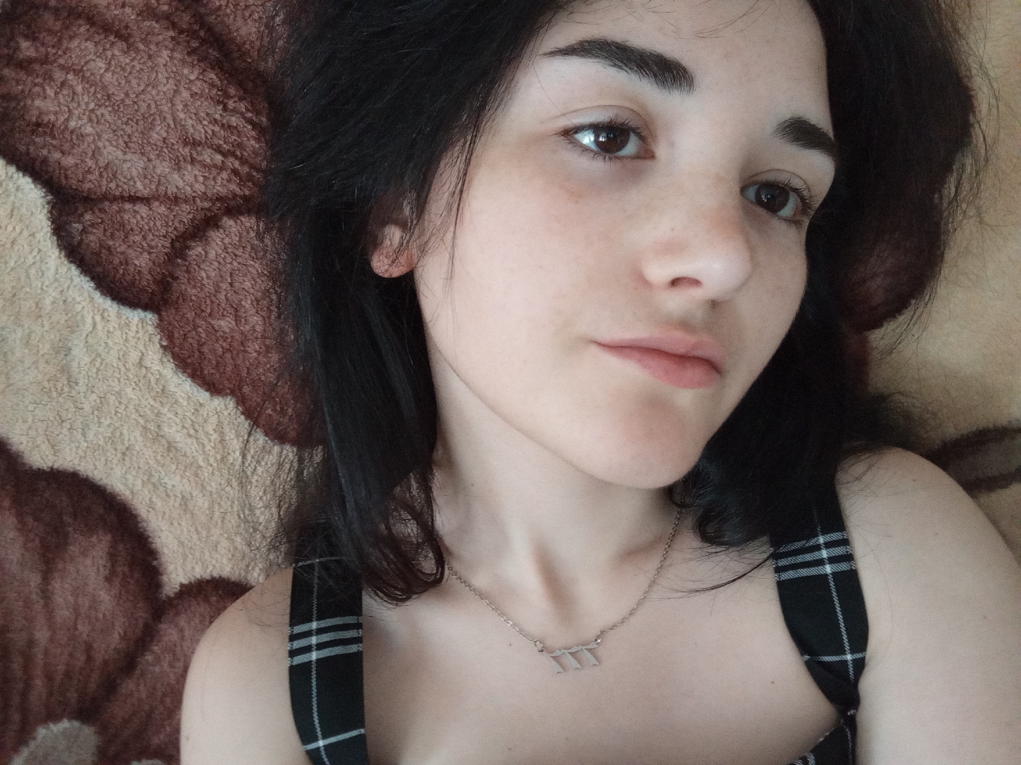 A close-up selfie shows a fair-skinned young woman with dark hair lying on a textured, brown and beige patterned surface, possibly a blanket or a rug. She is wearing a black and white plaid top with thin straps and a delicate silver necklace with a small pendant. Her facial features include well-defined eyebrows, brown eyes, and a slightly downturned mouth. Her complexion appears natural, with subtle freckles visible. The background is blurred, focusing attention on her face and upper body. There is no indication of location within the city of Unknown, Unknown.