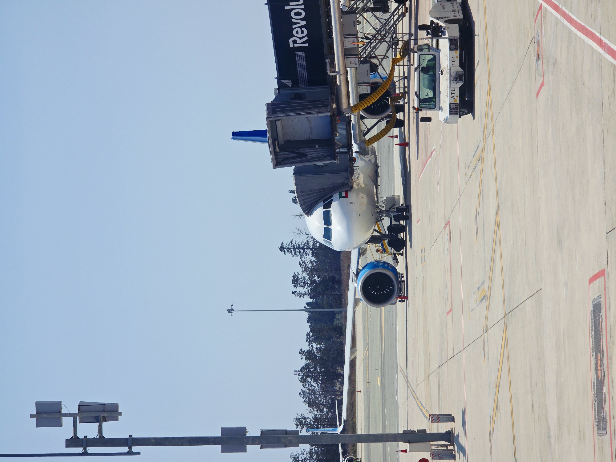 An airplane is parked at a gate at Larnaca International Airport, Cyprus. The airplane is white with blue accents on its tail and engines. A jet bridge is connected to the plane, and the word "Revolution" is visible on the jet bridge. A small white tow truck is parked in front of the jet bridge. In the background, there are trees and a clear blue sky. The airport tarmac is visible, with yellow and red markings on the ground. The scene is brightly lit by the sun, suggesting it is daytime.