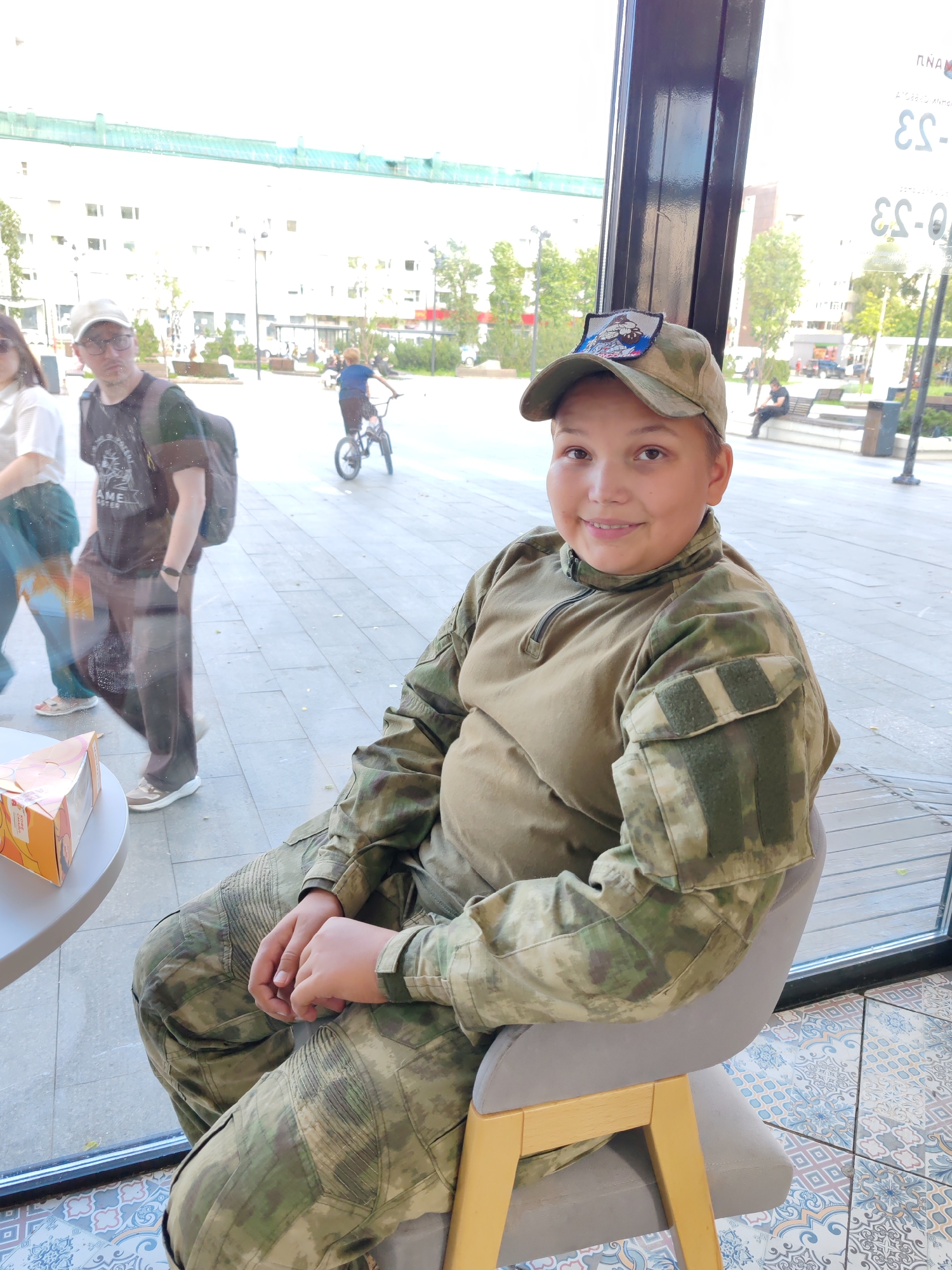 A young person wearing camouflage clothing and a baseball cap sits in a chair by a large window. The person is looking at the camera and smiling. Behind the person, through the window, a city street scene is visible, including buildings, trees, people walking, and a person riding a bicycle. The floor is tiled. The overall impression is an informal portrait taken in a public or semi-public space.
