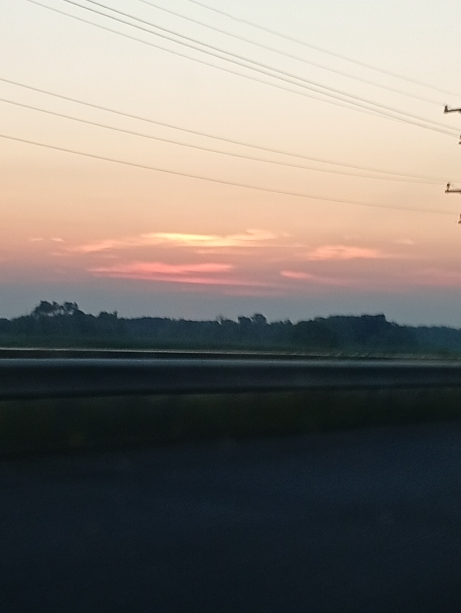 The image captures a wide shot of a sunset over a flat landscape. The sky is a gradient of pale yellow at the top, transitioning to soft pink and orange hues near the horizon. Several thin, dark wires run horizontally across the upper portion of the frame, suggesting the presence of power lines. Below the sky, a dark, silhouetted treeline stretches across the middle ground, with some indistinct shapes suggesting trees and possibly buildings. In the foreground, a dark, out-of-focus area dominates the bottom half of the image, likely representing a road or a dark surface. The scene appears to be an outdoor environment, possibly a roadside view, during twilight.