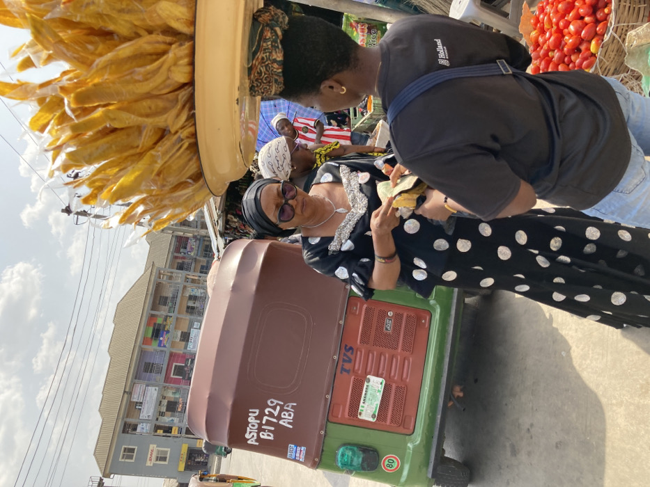 The image depicts a daytime street market scene in Aba, Nigeria. In the foreground, an adult female wearing a black top and long skirt with white polka dots, a black headwrap, and sunglasses is holding currency notes, engaging in a transaction with another individual. This second individual, facing away from the camera, wears a black t-shirt with "Holland" text and light blue jeans, and carries a large, light-yellow tray laden with individually plastic-wrapped yellow items (likely snacks) balanced on their head. A basket filled with red tomatoes is visible near their feet.
Immediately behind the polka-dotted woman, a green and brown motorized tricycle (keke napep) is parked. It bears the markings "ASTOPU B1729 ABA" in white paint on its rear, indicating its operational location. The tricycle is a "TVS" brand and displays a green Nigerian license plate reading "MSD ANAMBRA," along with a circular "80" speed limit sign. In the background, a multi-story building with balconies and various commercial signs, including one for "DHL," lines the street. Other individuals are visible in the mid-ground and background, contributing to the market atmosphere. Overhead power lines are visible against a clear, bright sky.