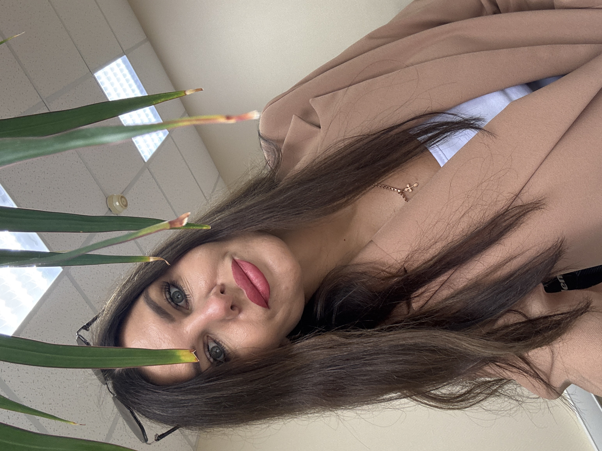 A woman with long, dark brown hair and green eyes is taking a selfie. She is wearing a light blue top and a tan blazer. She has on bright pink lipstick and a delicate gold necklace with a cross pendant. The background appears to be an office environment with a tiled ceiling and fluorescent lighting. The woman is looking directly at the camera with a slight smile. Green leaves hang in the foreground, partially obscuring her face and the background. The scene is likely indoors, possibly in a home office or a commercial workspace in the city of Chaikovsky, Russia.