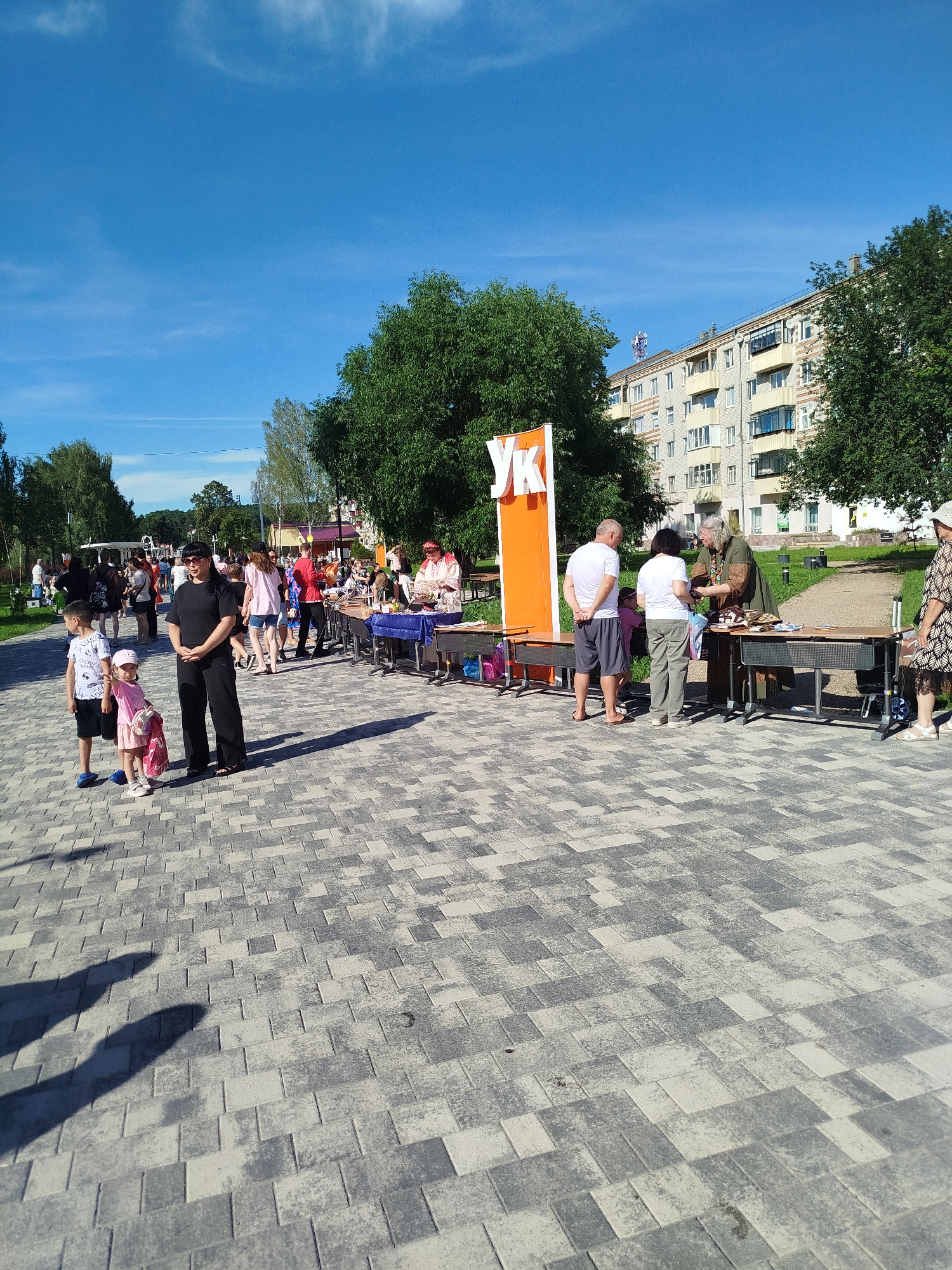 This image shows an outdoor event with multiple people gathered under a clear blue sky with scattered clouds. In the foreground, a paved walkway is visible, with a large building in the background. There are several tables set up, with people interacting around them, some appearing to be vendors or participants in an activity. A large orange sign with white letters "УК" is prominent on the left side of the image. Several trees and some grass are also present in the scene, suggesting a park or public space. The overall atmosphere appears to be a community gathering or festival.