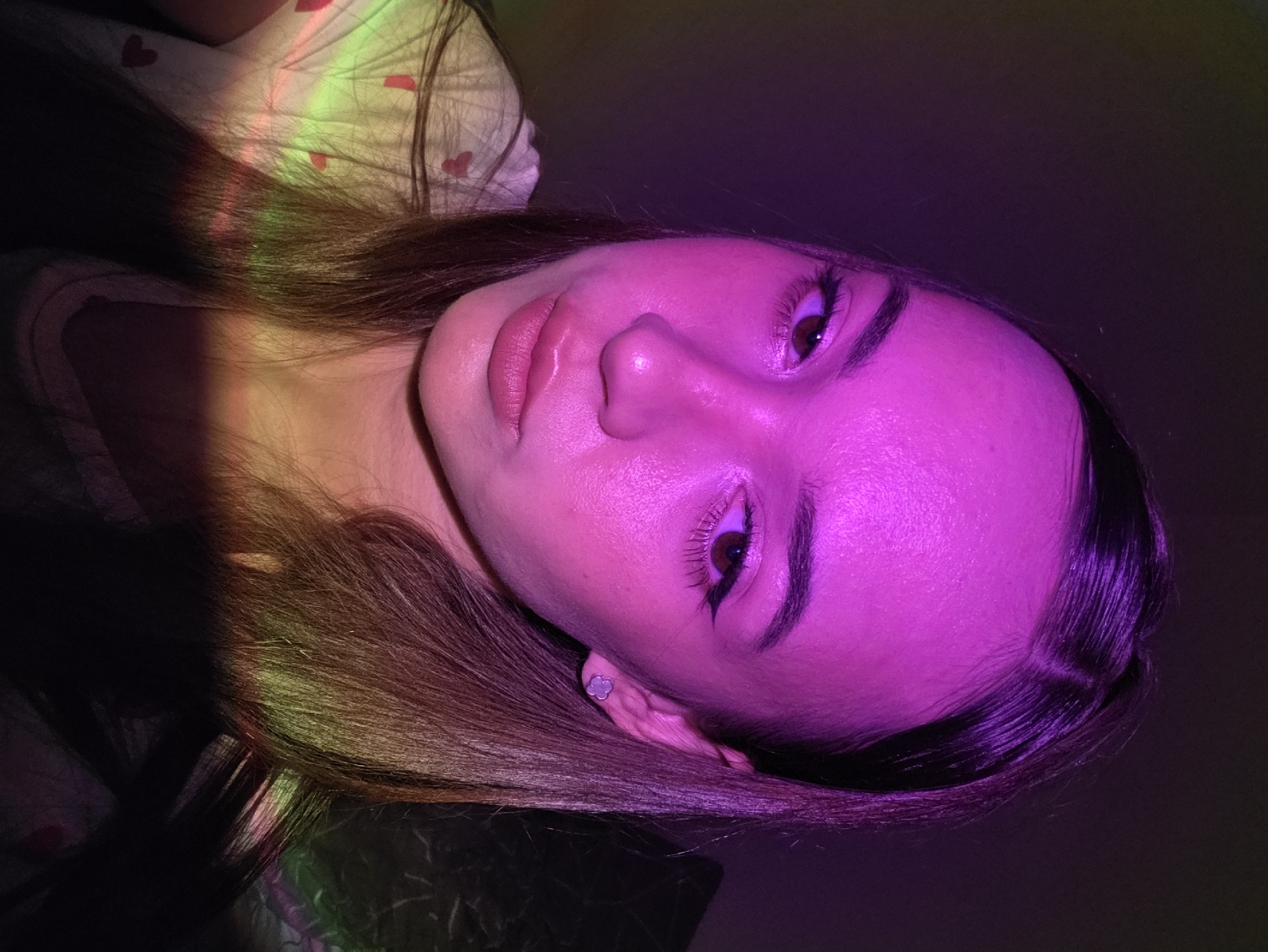 The image is a close-up selfie of a young woman taken in what appears to be a dimly lit indoor environment. She has fair skin, brown eyes, and dark brown hair, which is parted in the middle and falls past her shoulders. Her makeup includes winged eyeliner and lip gloss. She is wearing a light-colored t-shirt with small red heart patterns. The scene is illuminated by colored lights, casting purple and green hues on her face and clothing, creating a vibrant and atmospheric effect. The background is dark and indistinct. There is no information in the image to suggest a location in the city of Chaikovsky, Russia.