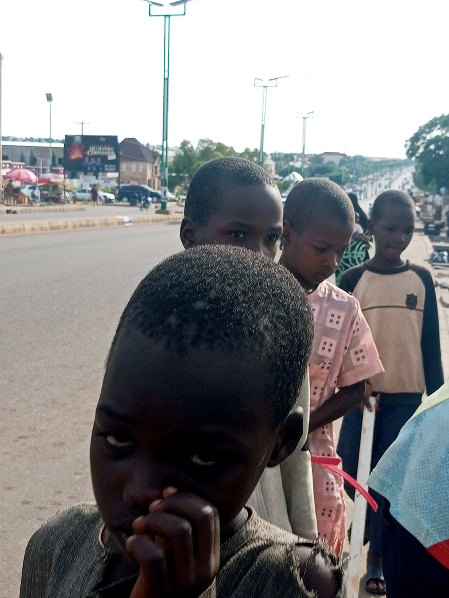 The image shows a group of four young children standing on a street in Tempure, Nigeria. The child in the foreground is looking down with their hand to their mouth. Behind them, three other children are also visible, one looking slightly to the side and two looking ahead. The children are dressed in casual clothing.
The scene is an outdoor environment with a paved road stretching into the distance, lined with trees and buildings. There are streetlights visible in the background, suggesting it might be daytime or early evening. Some vehicles and people can be seen further down the road, indicating a public area or thoroughfare. A billboard is visible on the left side of the image, with some text on it, though it is not clearly legible. The overall atmosphere appears to be casual and everyday. The children's expressions are difficult to discern clearly, but the child in the foreground seems pensive. The weather appears to be clear.