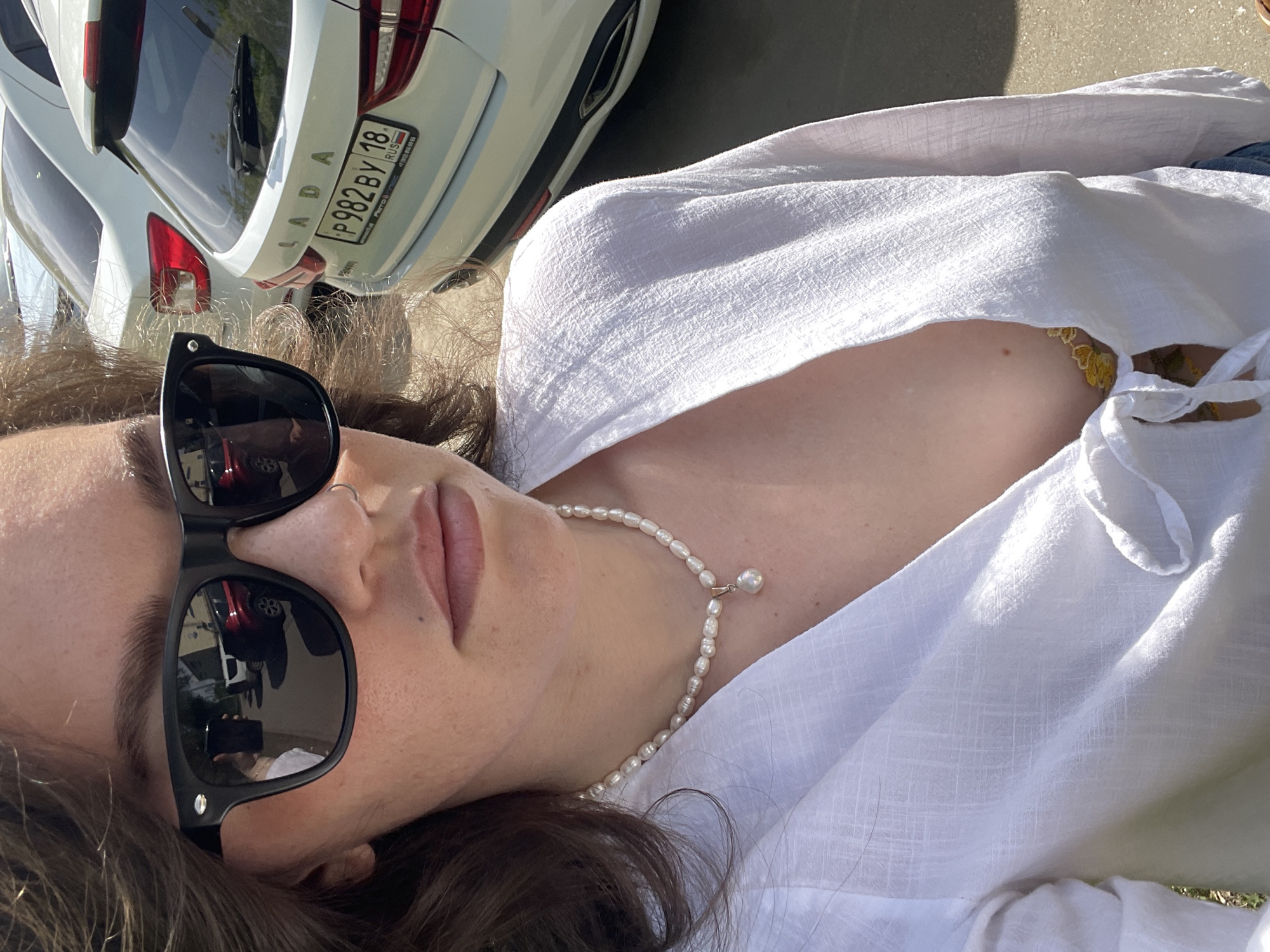 A person with sunglasses and a pearl necklace is lying down outdoors. The person is wearing a white shirt. Behind them is a white Lada car with the license plate P982BY 18 RUS. Based on the license plate, the location is Izhevsk, Russia.