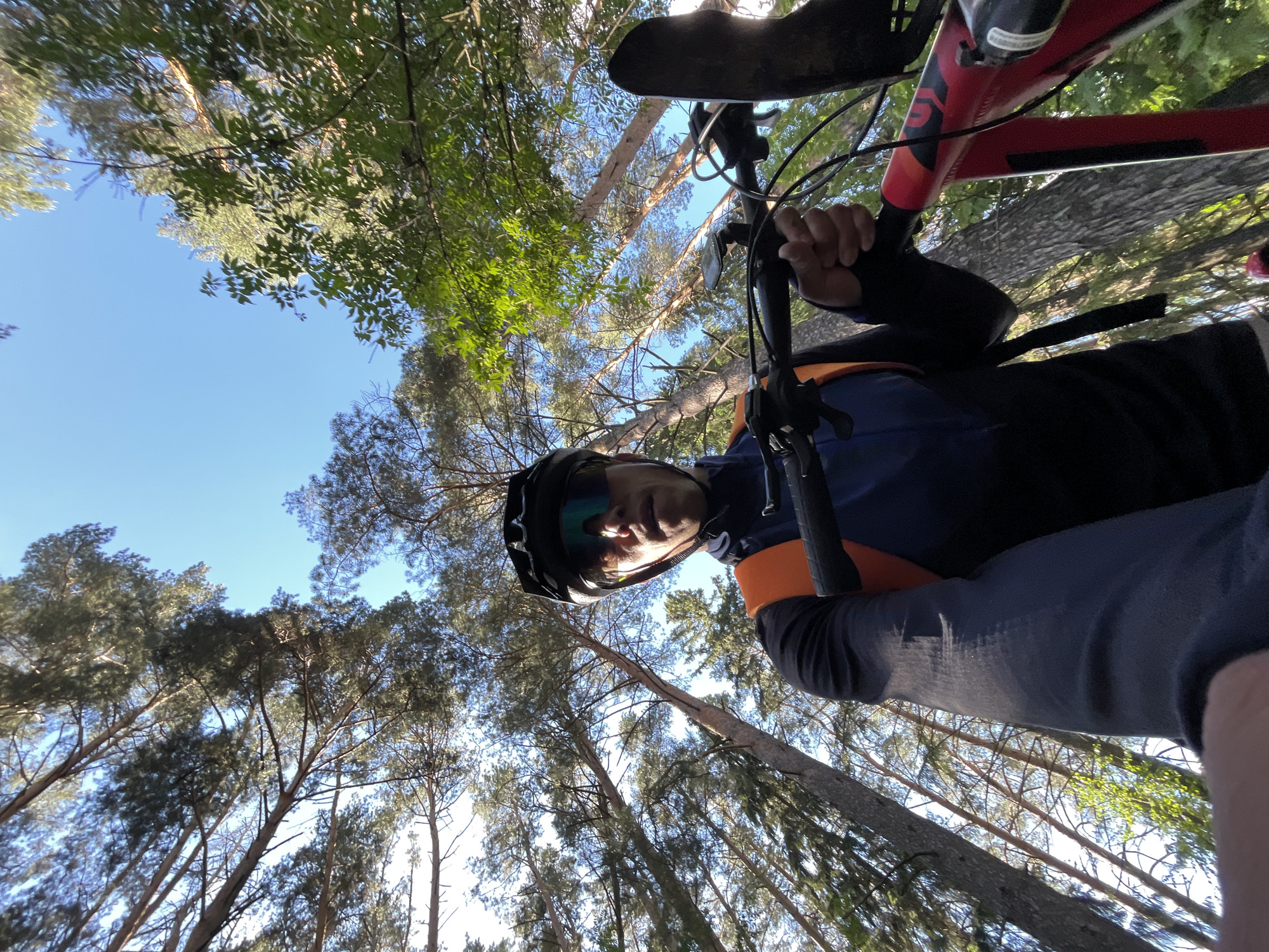 A low-angle shot shows a person wearing a dark-colored helmet and sunglasses, holding the handlebars of a red and black bicycle. The person is wearing a long-sleeved cycling jersey with a dark blue body and orange shoulders. The background consists of tall trees, with branches reaching toward a bright blue sky. The location is likely a wooded area in Chaikovsky, Russia.