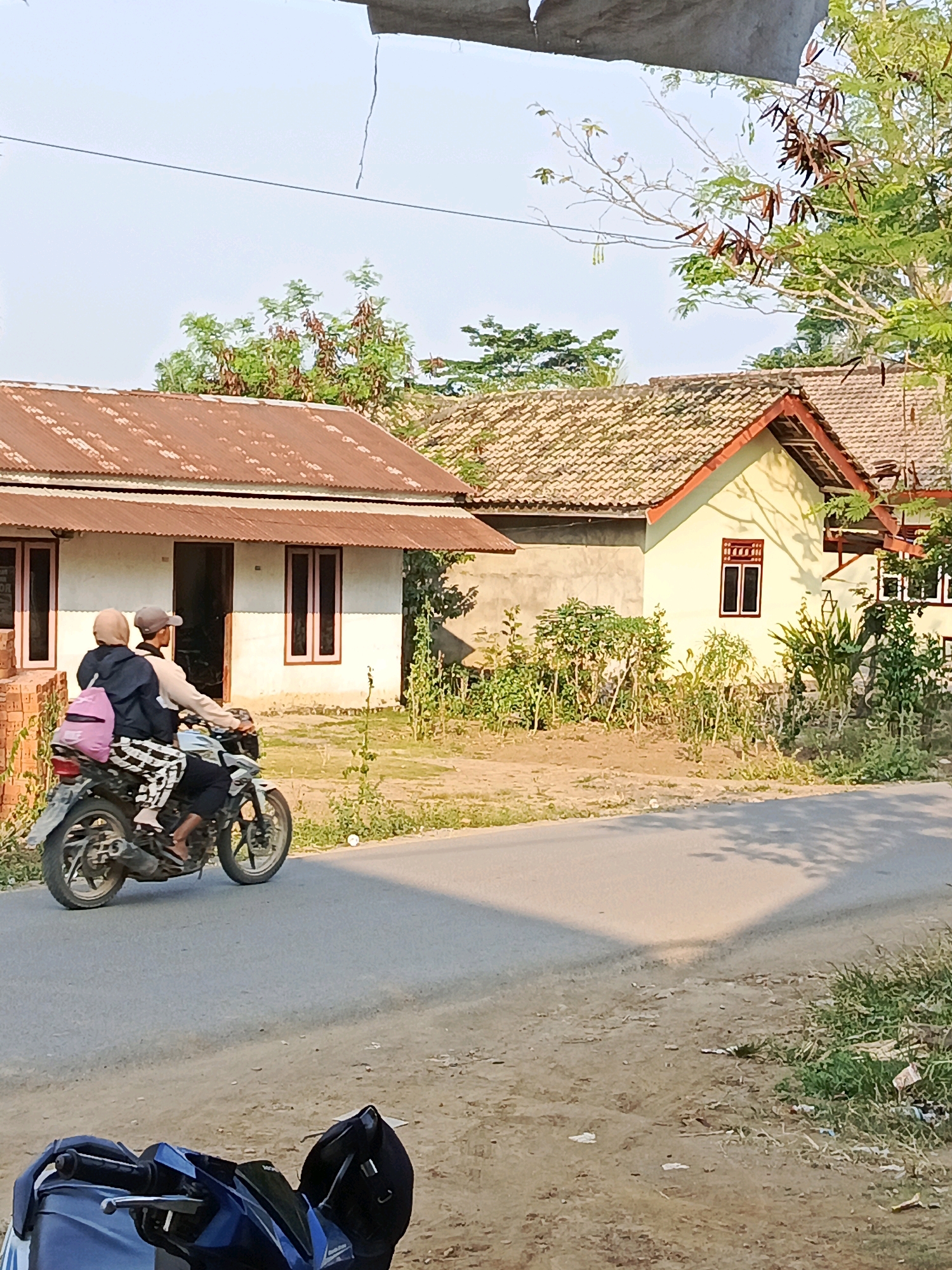 Two individuals are riding a motorcycle on a paved road in front of a residential area in Megang Sakti I, Indonesia. The rider, wearing a cap and a light-colored shirt, is looking forward. The passenger, wearing a headscarf and a dark jacket, is facing away from the camera and has a pink backpack on their lap. They are both wearing patterned pants. The motorcycle is white with black accents. The background features two houses with corrugated metal roofs, one with a rust-colored roof and the other with a lighter, tile-like roof. Vegetation, including trees and bushes, is present around the houses and along the roadside. The sky is clear and blue. A portion of another motorcycle is visible in the foreground, on the right side of the image.