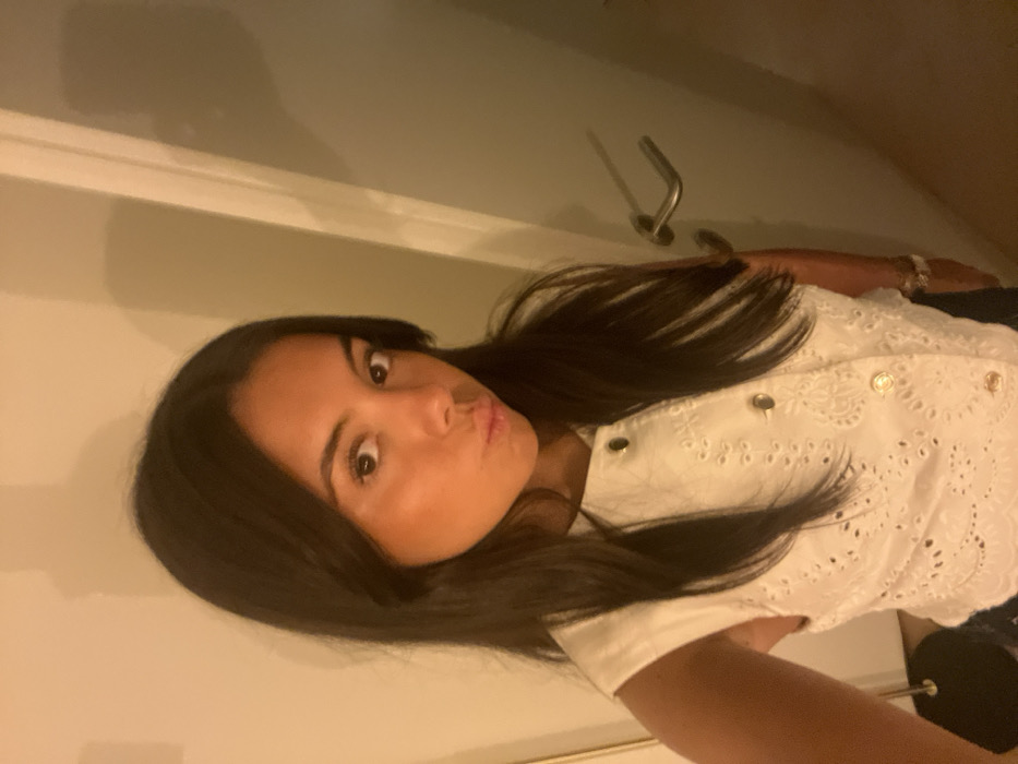 An indoor, waist-up self-portrait depicts a young adult female, standing with her upper body angled slightly to the right, looking directly at the camera. She has long, dark hair, a tan complexion, and brown eyes. Her lips are puckered in a "kiss" or "duck face" expression. She wears a white sleeveless blouse with an eyelet embroidery pattern and a vertical row of gold-toned buttons down the front. A silver-colored watch is visible on her left wrist. The background features a plain, light-colored wall to the left and a light-colored door with a horizontal silver-toned handle to the right. A dark, thin, vertical object is partially visible at the bottom left. The environment is illuminated by warm-toned artificial light, casting subtle shadows. The image lacks specific identifying features to determine a precise location within Lordelo do Ouro e Massarelos, Portugal, beyond being an interior space.
