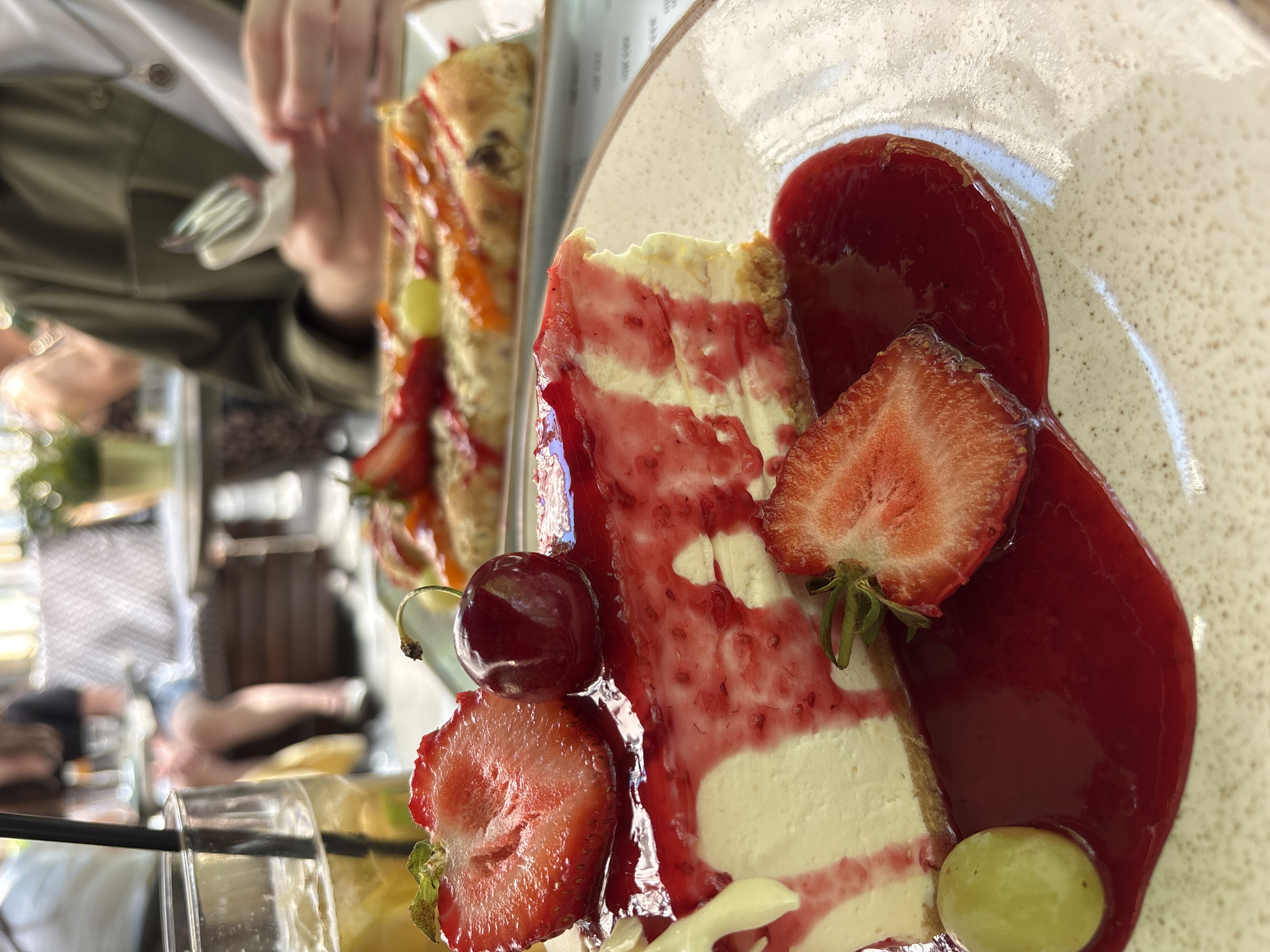 An outdoor dining scene in Belgrade, Serbia, features a close-up of a slice of cheesecake on a speckled plate. The cheesecake is topped with red berry sauce and garnished with a halved strawberry, a whole cherry, and a green grape. In the background, a person is visible holding a fork and napkin, and another person is seated at a table with a drink. The setting appears to be a restaurant or cafe with blurred foliage and outdoor seating.