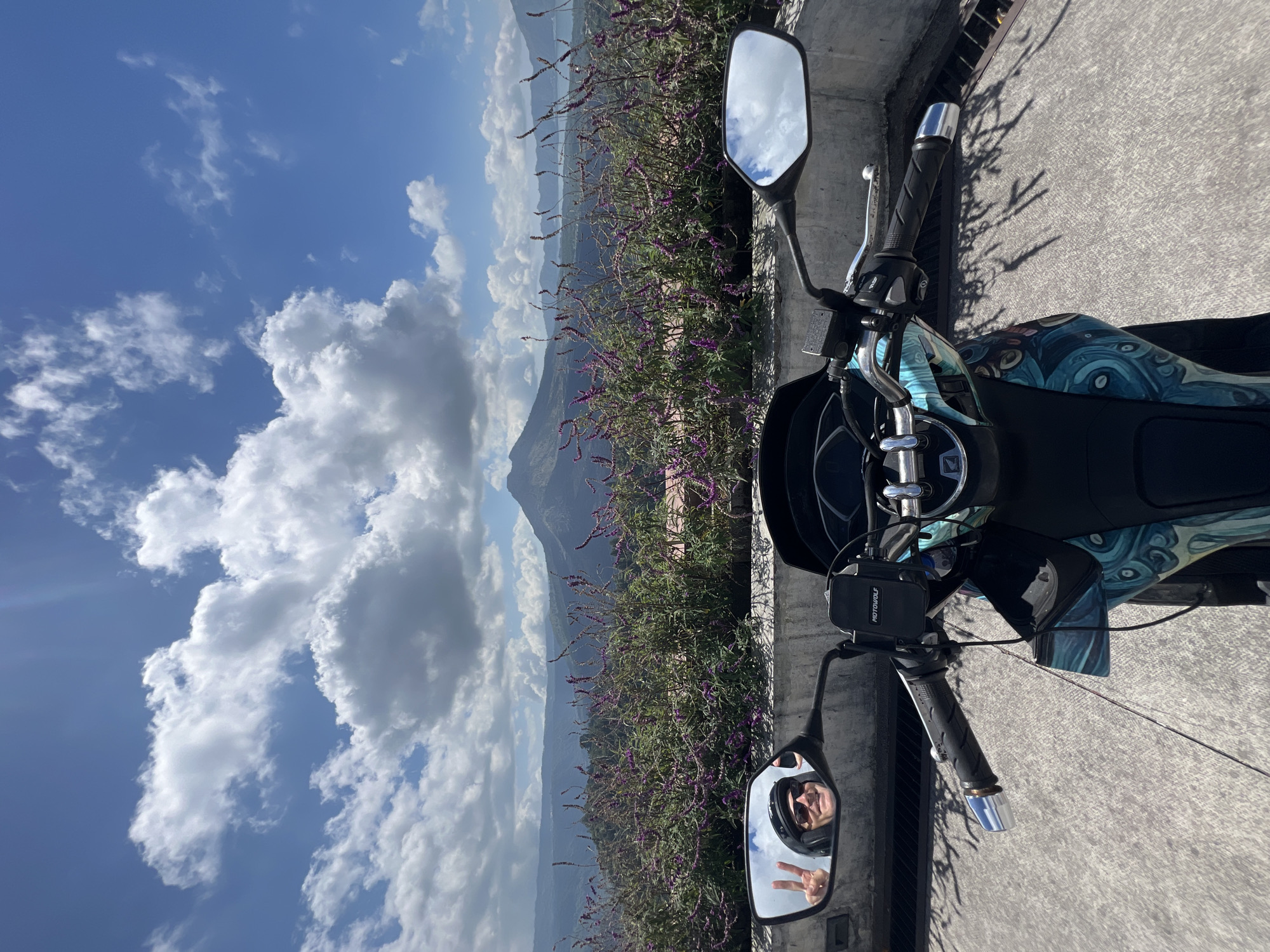A first-person perspective shot of a person on a scooter in Kintamani, Indonesia, on a sunny day. The rider, wearing a helmet and sunglasses, smiles and gives a peace sign in the left mirror. The scooter's handlebars, mirrors, and front panel are visible. Beyond the scooter, a line of purple flowers sits atop a concrete barrier, with a mountain and lake in the background. The sky is blue with white clouds.