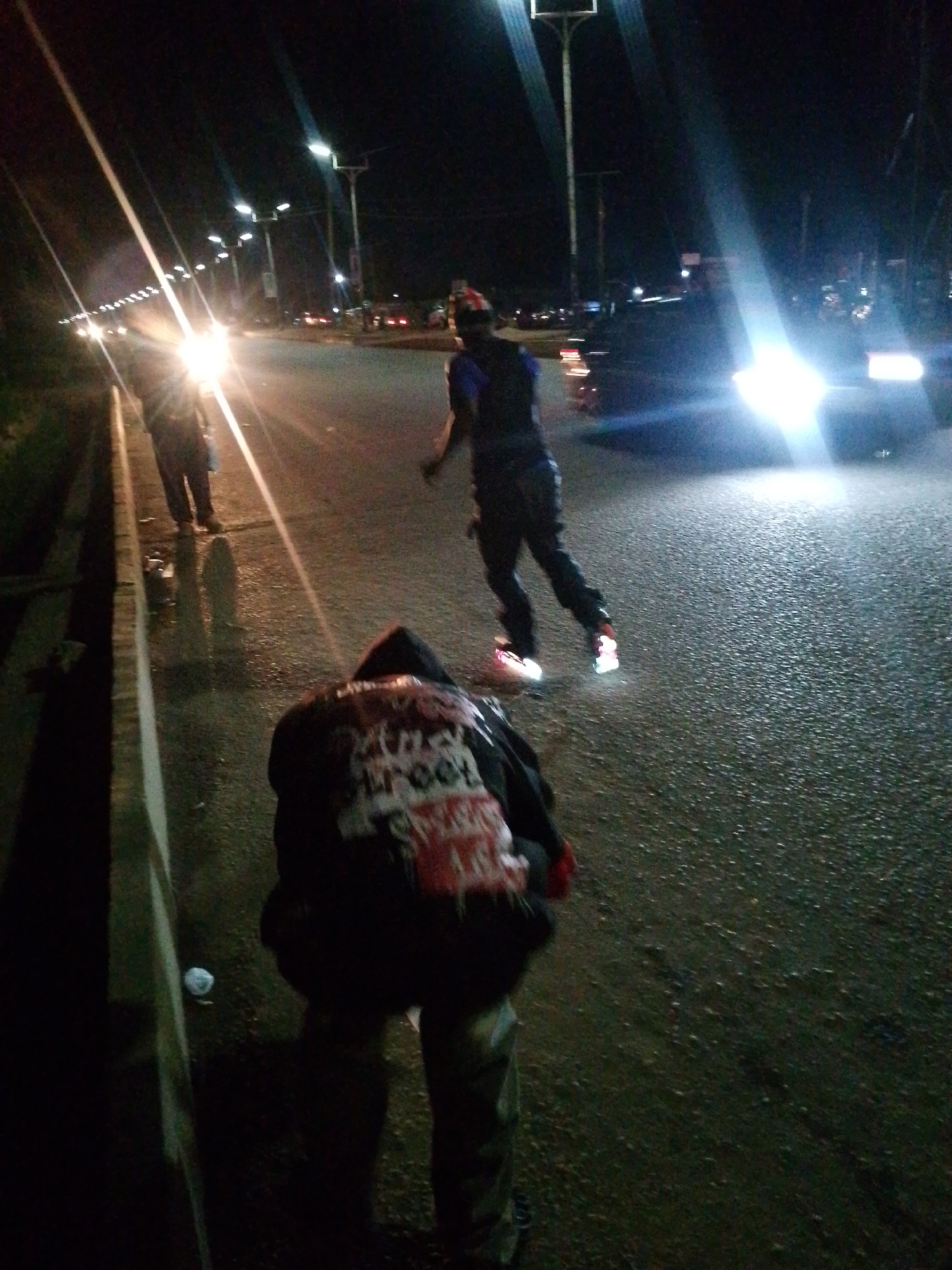 The image depicts a nighttime street scene in Bukuru, Nigeria, with the road illuminated by streetlights and vehicle headlights. In the foreground, a person wearing a hooded jacket with a red and white design on the back is partially visible, facing away from the camera. Further down the road, another individual in a blue shirt and dark vest is walking. In the background, several vehicles, including cars and possibly a bus, are present, their headlights contributing to the overall illumination. The road appears wet, suggesting recent rain, and is lined with a curb on the left side. The environment appears to be an urban or semi-urban setting, with buildings and streetlights visible in the distance. The overall impression is that of a busy street at night, possibly a thoroughfare within the city.