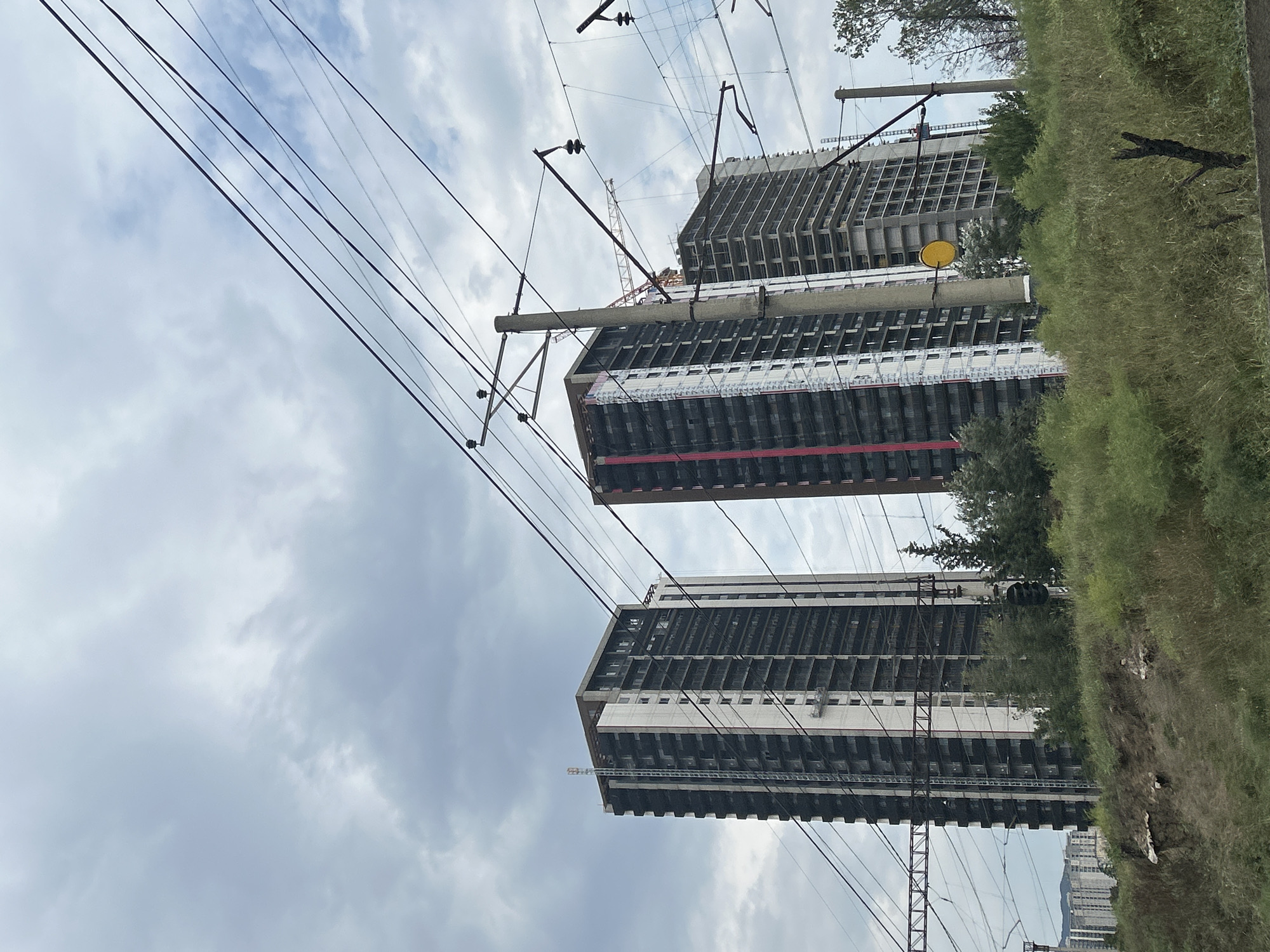 This is a photograph of construction sites with several high-rise buildings under development, located in Tbilisi, Georgia. The image captures a cloudy sky with multiple power lines crisscrossing the frame. In the foreground, a vegetated embankment with some dry branches is visible. The main subjects are three unfinished apartment buildings, two of which are partially covered in construction sheeting and scaffolding. One of the buildings features a prominent red stripe running vertically. A construction crane is visible behind one of the buildings. In the background, other buildings are visible, suggesting a developing urban area. Electrical poles and wires are prominent throughout the scene, indicating proximity to transportation infrastructure.