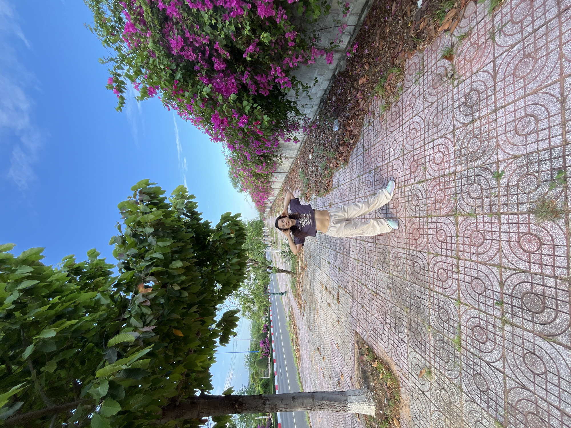 In Phu Thuy Ward, Vietnam, under a clear blue sky with sparse white clouds, a young woman lies on a patterned tiled sidewalk. She is positioned near a wall covered with vibrant purple bougainvillea flowers, which cascade down, creating a colorful border along the walkway. To her left, large green trees with lush foliage dominate the foreground. The sidewalk itself features a repeating circular and grid pattern in red and grey tiles, with patches of grass and fallen leaves interspersed.
The woman is lying on her back, looking up and smiling, with her hands behind her head. She wears a dark t-shirt that exposes her midriff and light-colored loose-fitting pants. Her light blue sneakers are visible on her feet. The scene appears to be set during the daytime, likely morning or afternoon, given the bright sunlight casting shadows.
Beyond the sidewalk and bougainvillea, a road is visible with traffic markings and a few trees lining its edges, suggesting a street or pathway in a semi-urban or park-like setting. The overall atmosphere is relaxed and cheerful, with the woman enjoying a moment of leisure outdoors amidst the natural beauty of the flowering plants and trees.