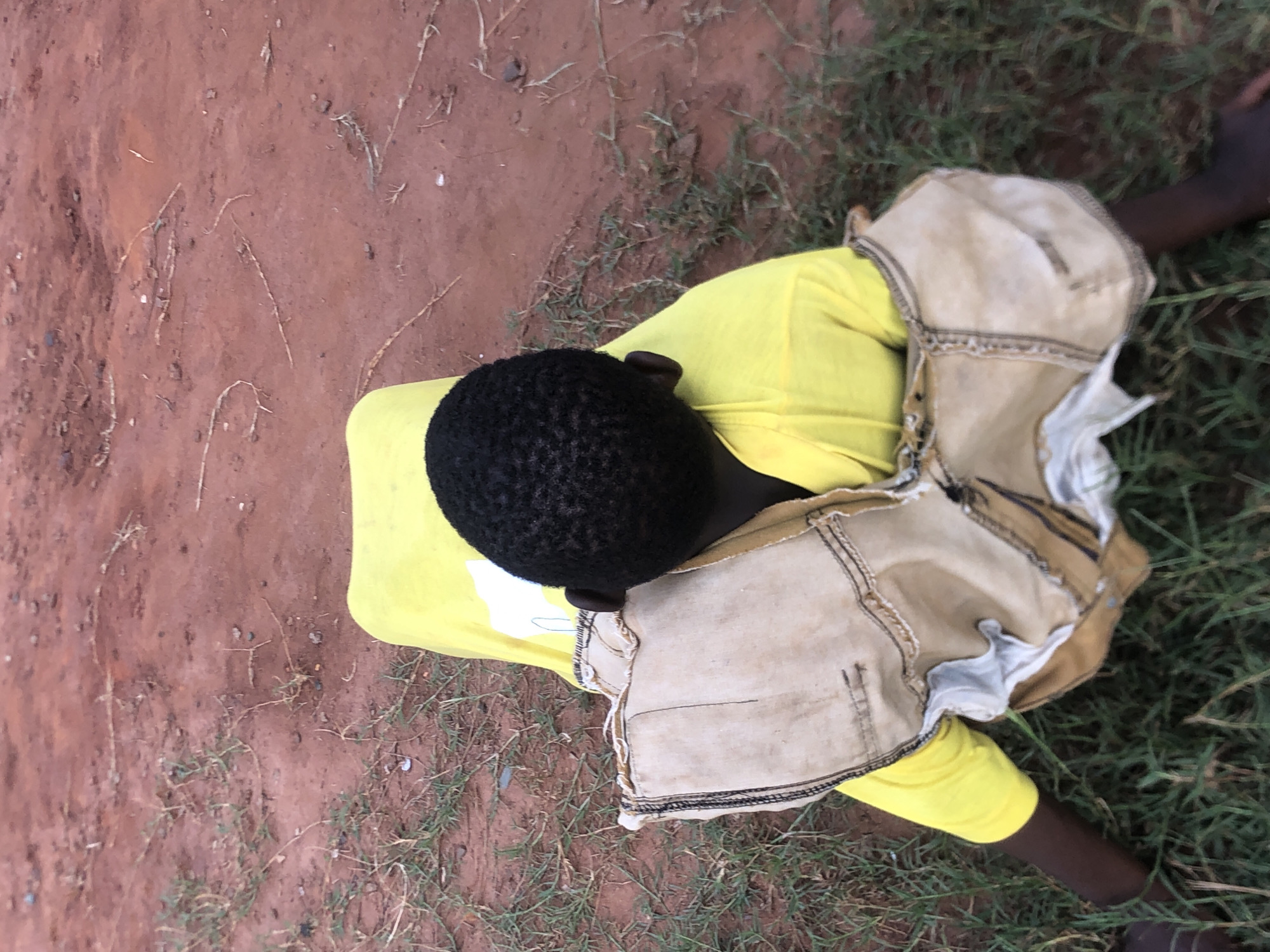 An overhead shot captures a person lying or sitting on the reddish-brown soil in Ukwu-Abwa, Nigeria. The individual, likely a child or young person, is viewed from directly above, with their head positioned towards the upper left of the frame. They are wearing a bright yellow long-sleeved shirt. Over the yellow shirt, a light-colored, possibly beige or tan, distressed garment with prominent stitching and frayed edges covers their lower torso and legs, resembling worn shorts or a skirt.
The person's head, covered in short, tightly curled black hair, is visible. Their left ear is partially seen. A dark-skinned arm extends outwards towards the right, resting on patches of green grass interspersed with the soil. The ground is a mix of bare reddish-brown earth and sparse green foliage, characteristic of an outdoor, possibly rural, setting. Small stones and dry plant matter are scattered across the soil. The lighting suggests it is daytime, though the exact time is not discernible. No specific activity beyond resting or lying on the ground can be clearly identified, and emotions are not visible. There is a small white rectangular patch on the yellow shirt, but no legible text is visible on it or elsewhere in the image.