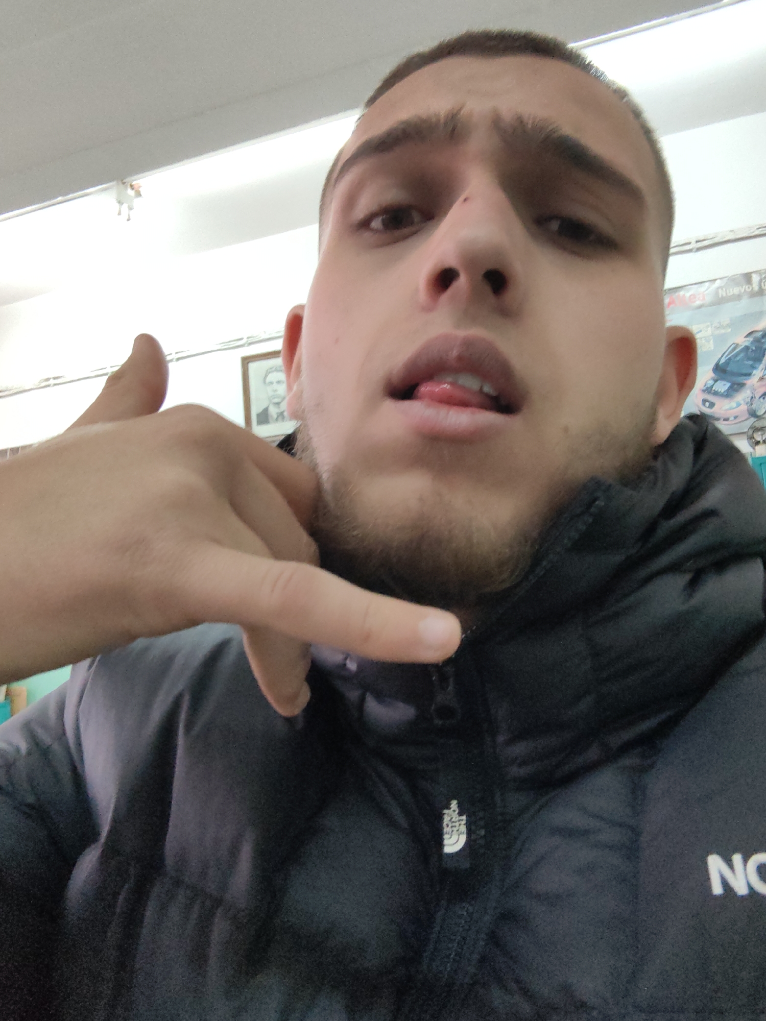 A close-up photo depicts a light-skinned young man with short, buzzed hair, a slight beard, and a small red mark near his nose. He is wearing a dark-colored puffer jacket with the "The North Face" logo visible on the front. He is making a "call me" gesture with his hand. The background is blurry, but a framed picture of a man and a poster featuring a car are visible, suggesting the location could be inside a building. The location in the city of Sofia, Bulgaria, cannot be determined from the image.